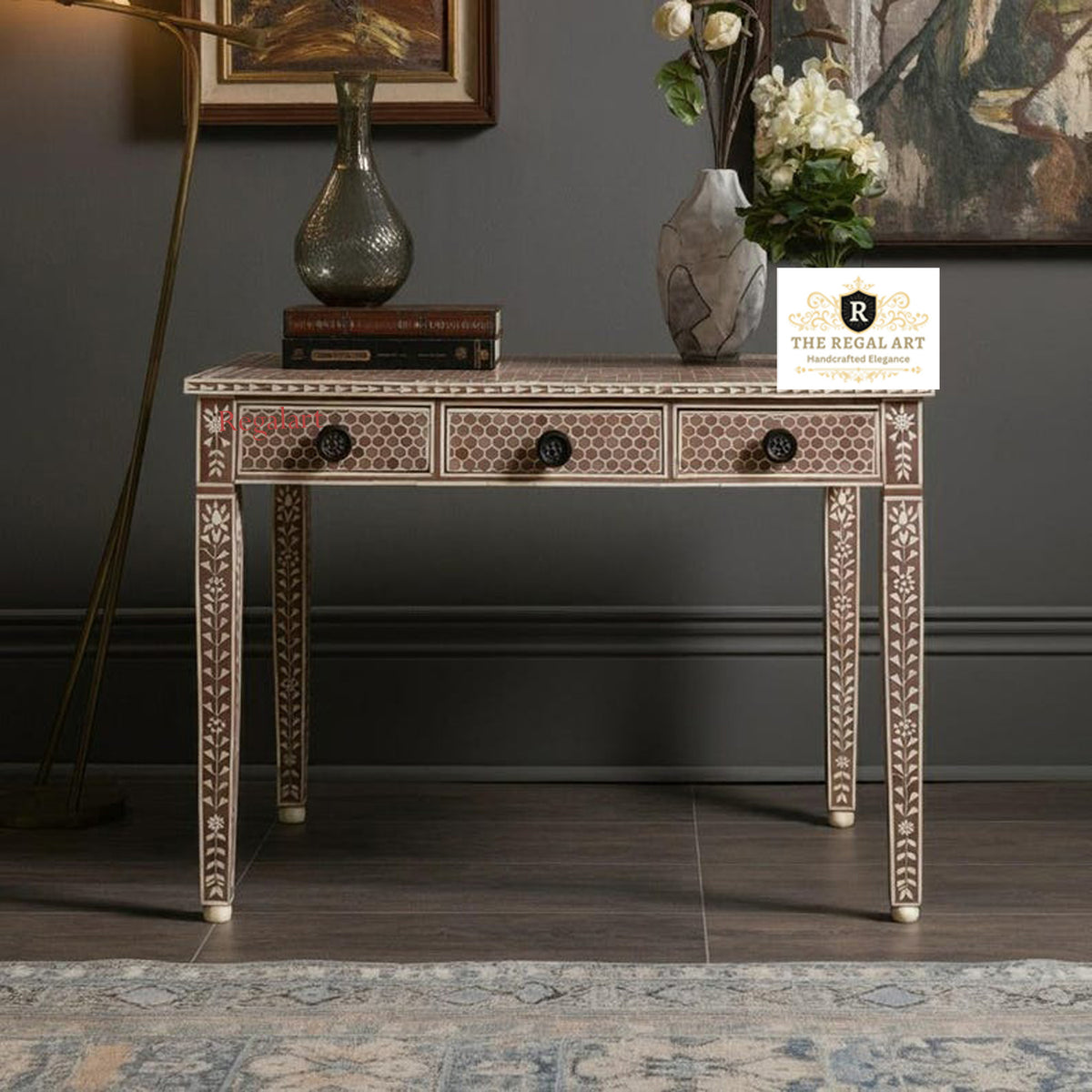 A soft pink desk with leaf pattern bone inlay work and three drawers, placed in a room with decorative items on top and a flower arrangement in a vase in the background.