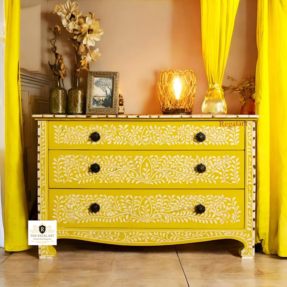 A yellow floral dresser with three drawers, featuring bone inlay work, placed against a backdrop of yellow curtains and decorative items.