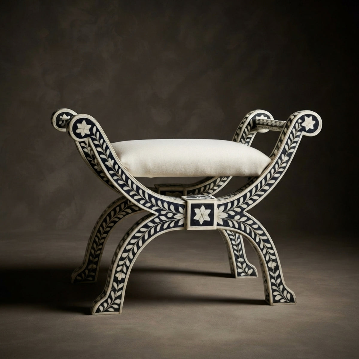 An ornate bench with a curved bone-in-wood frame featuring dark blue inlay and a white cushion, set against a dark textured studio background.