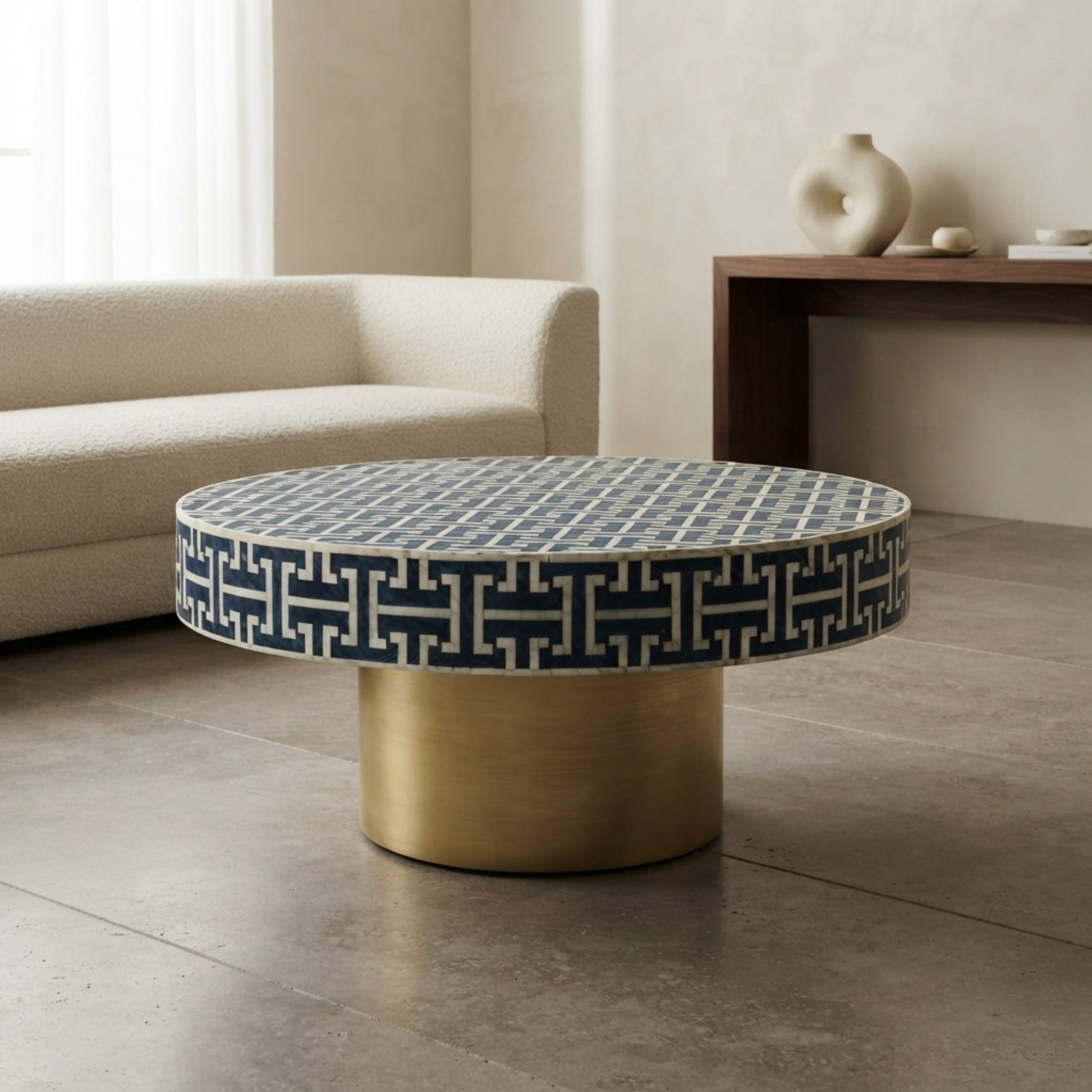 A round coffee table with a navy blue and white geometric patterned top and a brushed gold cylindrical base, set in a modern living room next to a cream sofa.