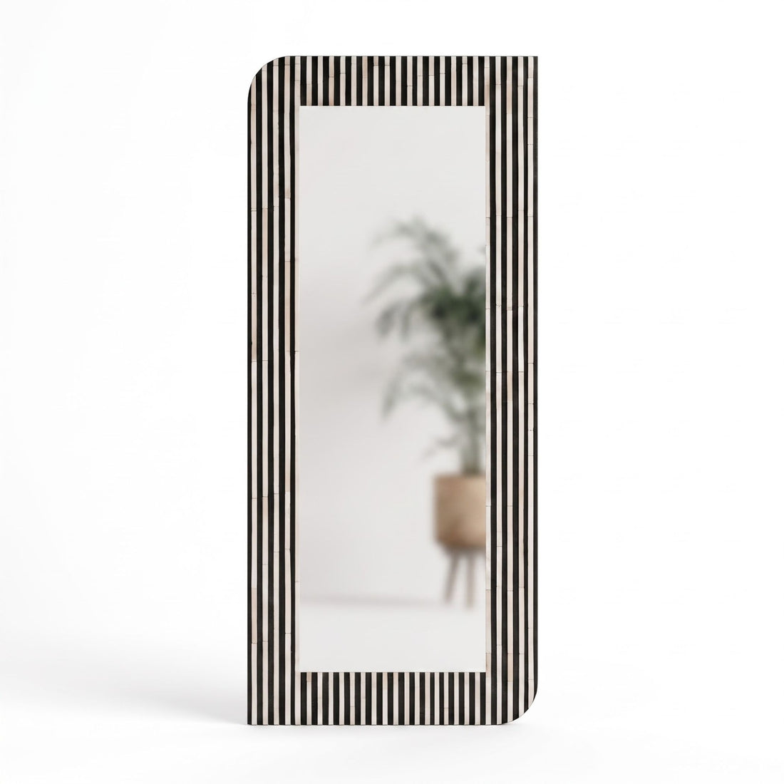 A tall rectangular floor mirror with a black and white vertically striped frame, styled in a minimalist beige-toned room with a marble console table and a potted olive tree reflected in the glass.