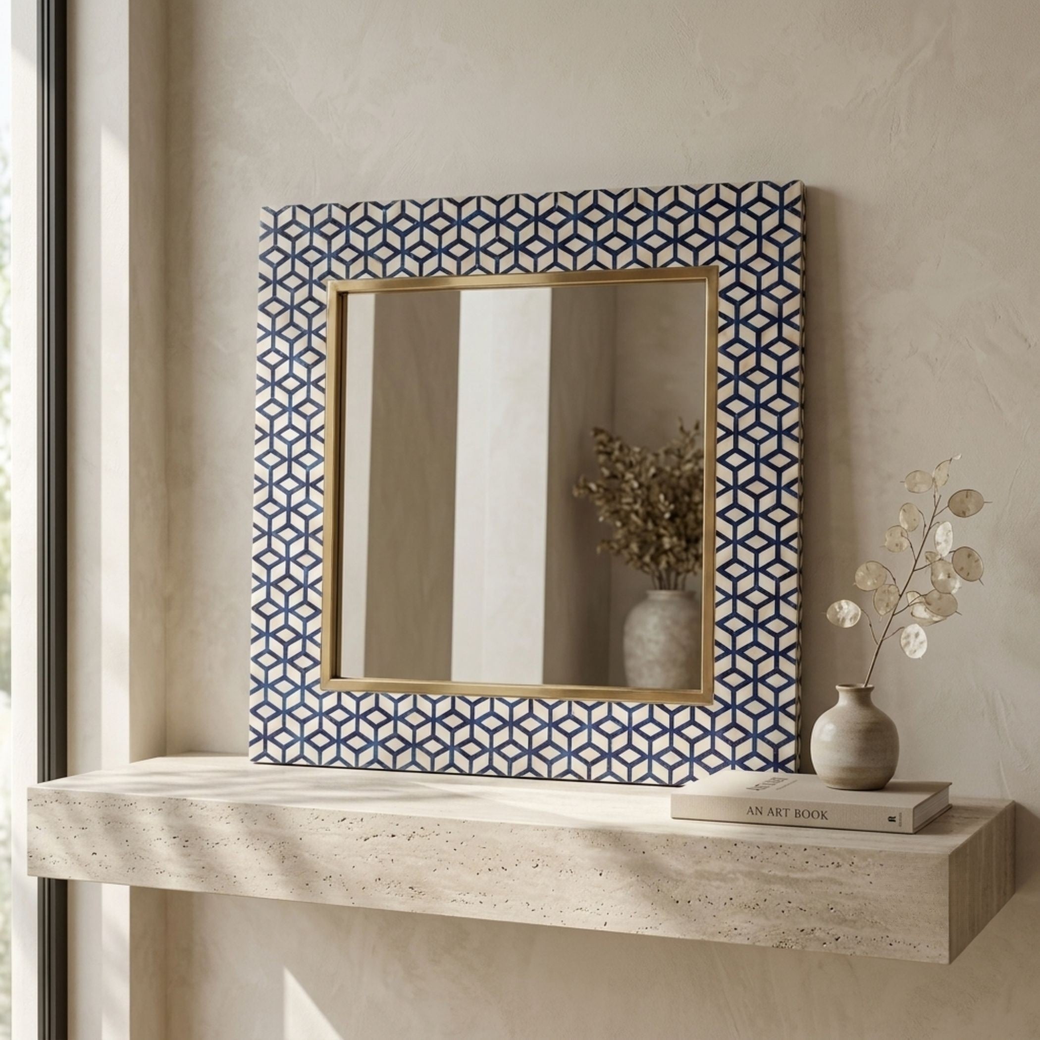 A rectangular wall mirror with a geometric blue and white patterned frame and a gold inner border, displayed on a light stone shelf against a textured beige wall with decorative vases.