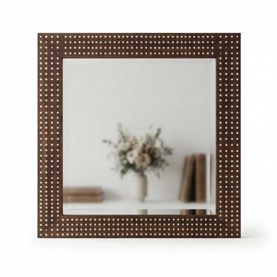 A large wall mirror with a dark brown frame studded with small metallic dots, shown hanging above a console table in a styled interior. The reflection and room setting include a vase with branches, books, a lit table lamp, and part of a textured chair.