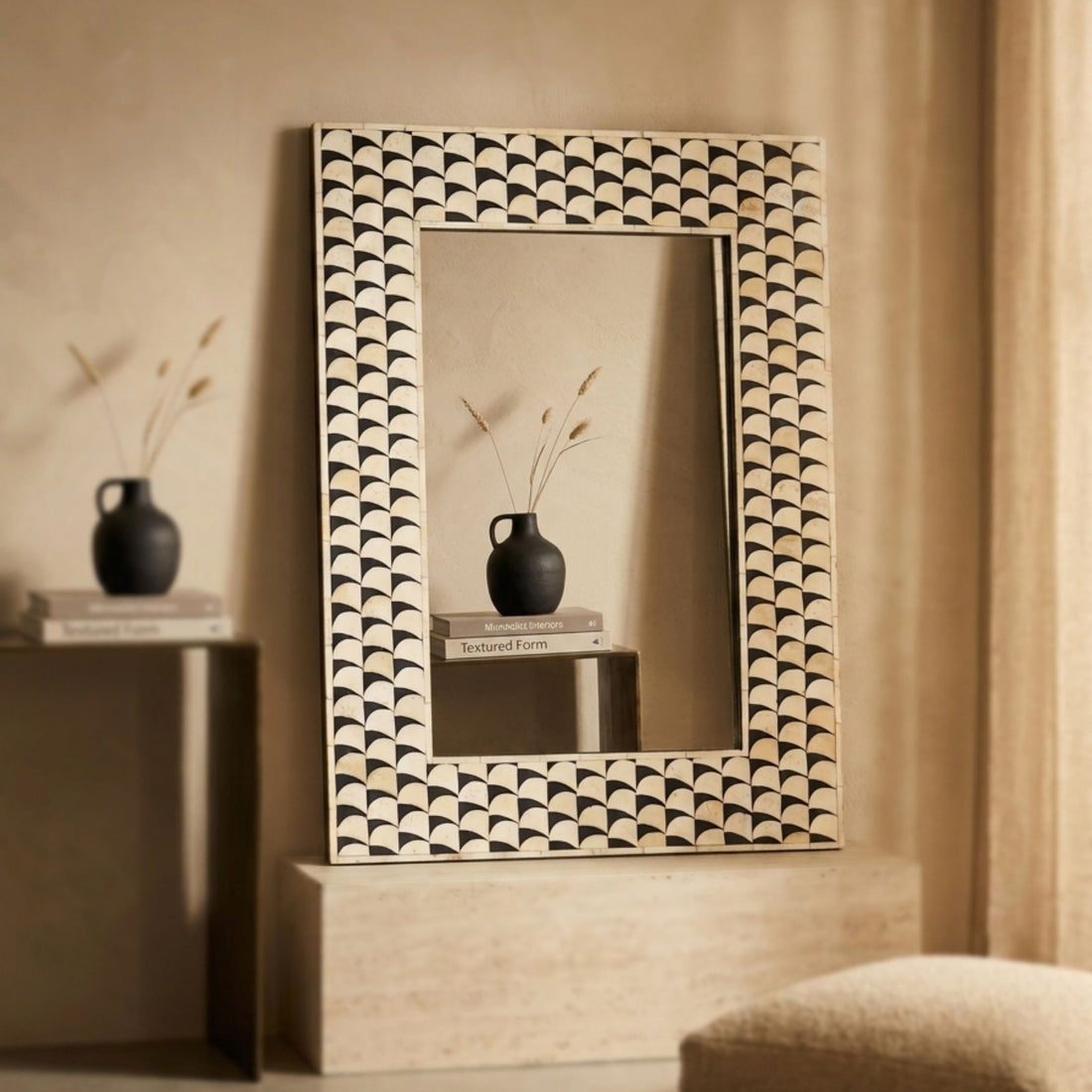 A rectangular wall mirror with an intricate black and beige scalloped geometric frame, styled on a light stone pedestal in a warm, neutral-toned room.