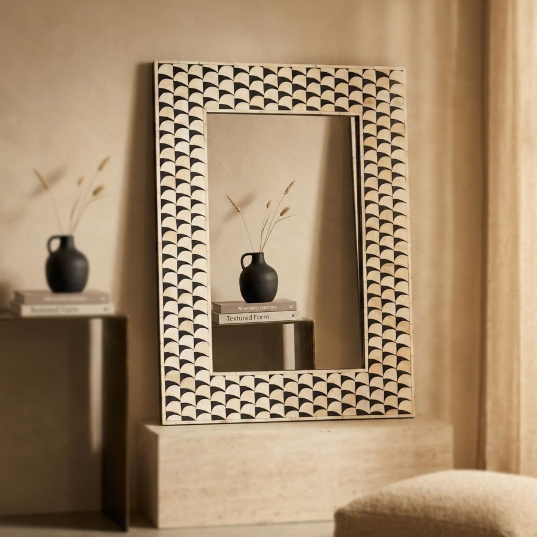 A rectangular wall mirror with an intricate black and beige scalloped geometric frame, styled on a light stone pedestal in a warm, neutral-toned room.