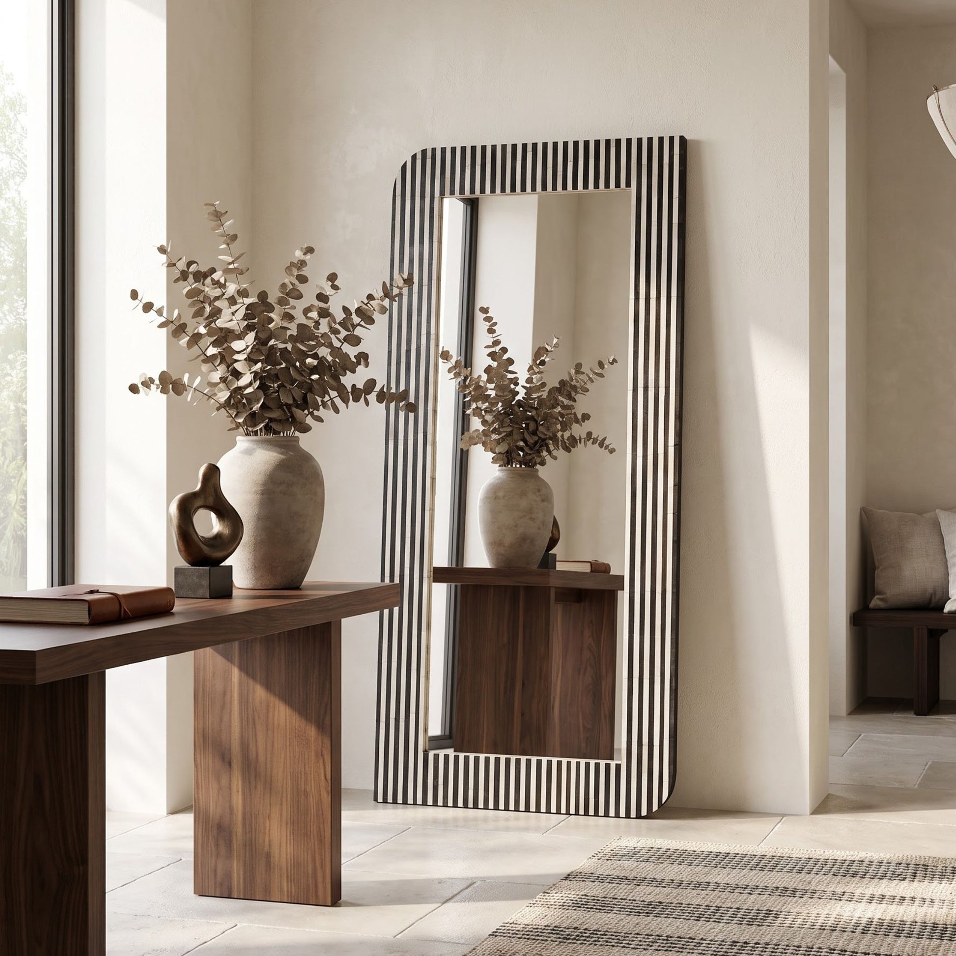 A tall rectangular floor mirror with a black and white vertically striped frame stands in a styled interior room next to a wooden console table. The mirror reflects a matching ceramic vase with dried botanicals, and the room has warm neutral walls, light stone flooring, and a woven rug.