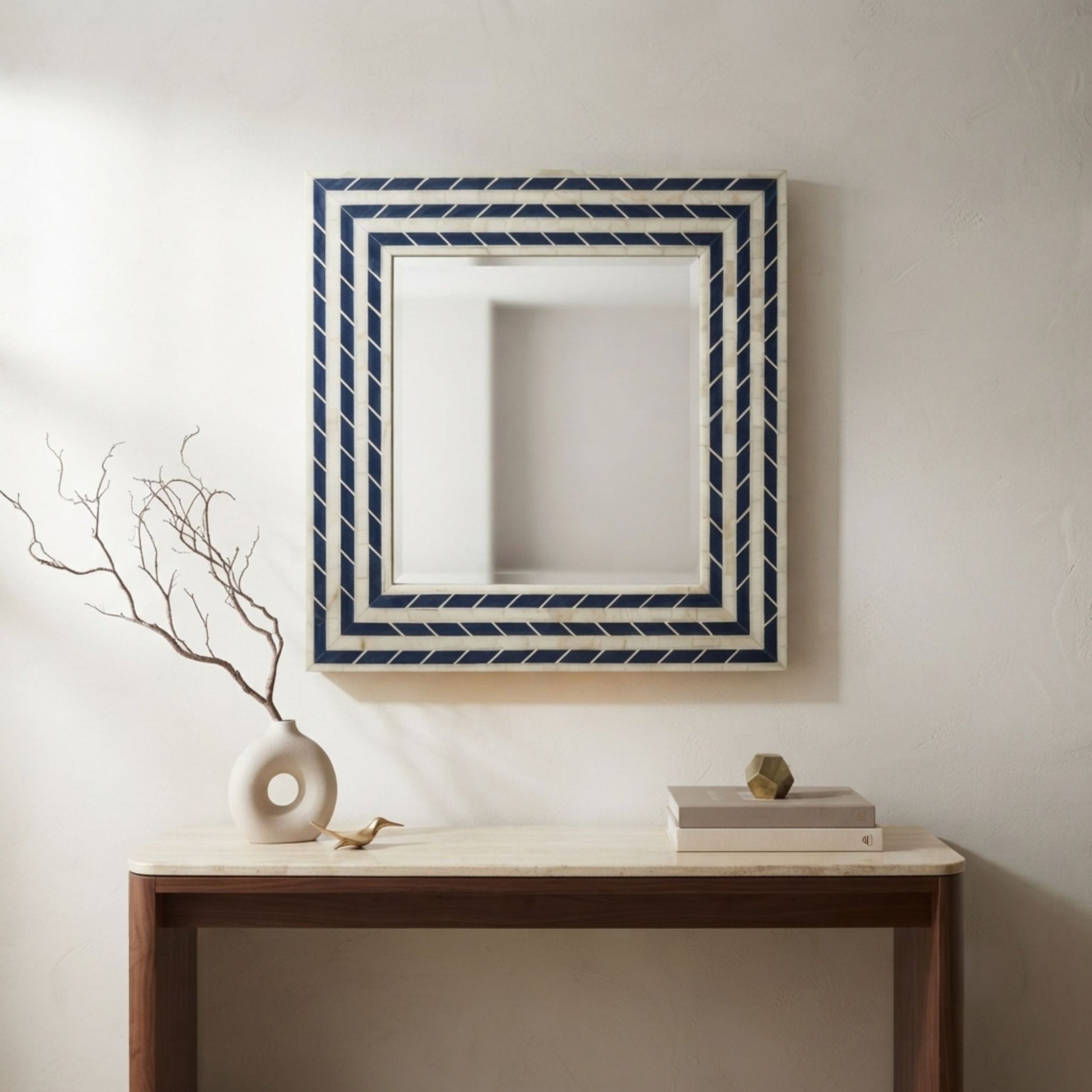 A square decorative wall mirror with a navy blue and white geometric patterned frame, hanging on a textured off-white wall above a wooden console table with a vase and books.