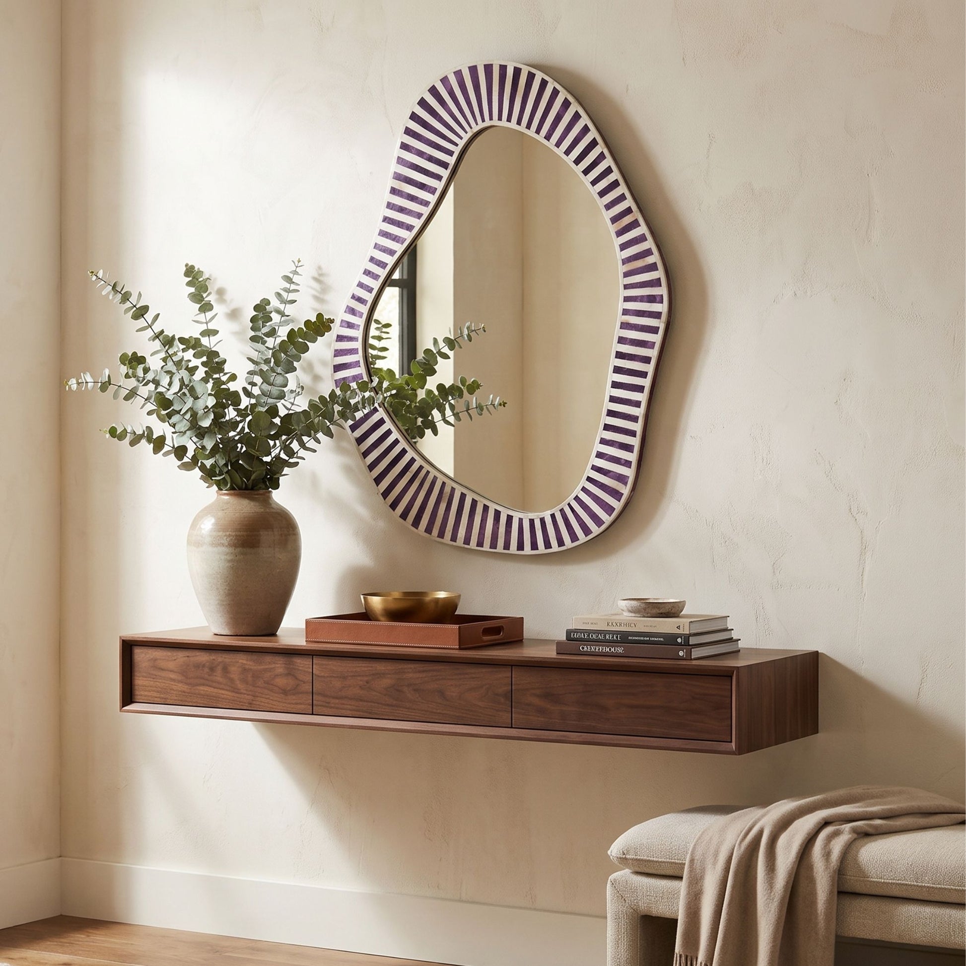 An organic-shaped wall mirror with a purple and white striped frame hangs on a beige wall above a floating dark wood console table. The scene is styled with a ceramic vase of greenery, books, and a bench with a draped throw blanket.