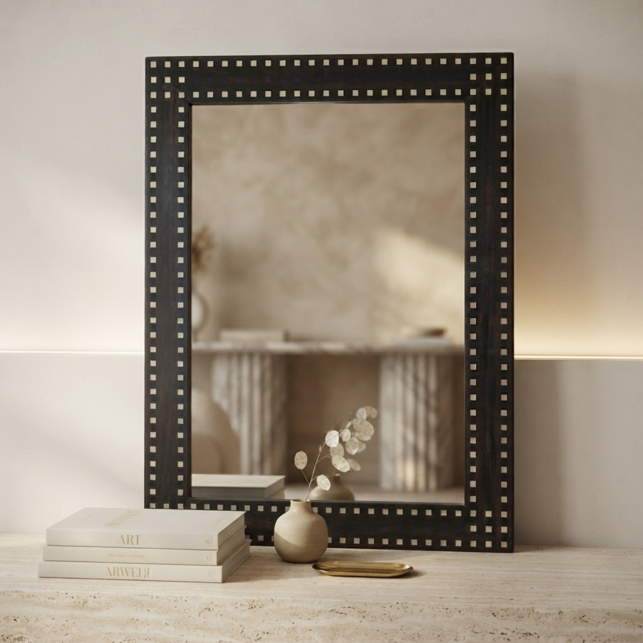 A rectangular black-framed mirror with a decorative border of small metallic studs, styled on a light stone surface with stacked books, a small vase, and dried stems against a warm beige wall.