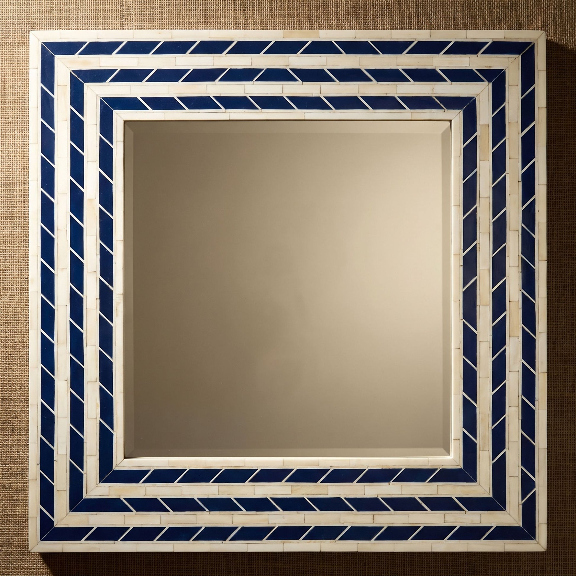 Square decorative wall mirror with a geometric mosaic border in navy blue and white tile-like pieces, shown against a textured beige background.