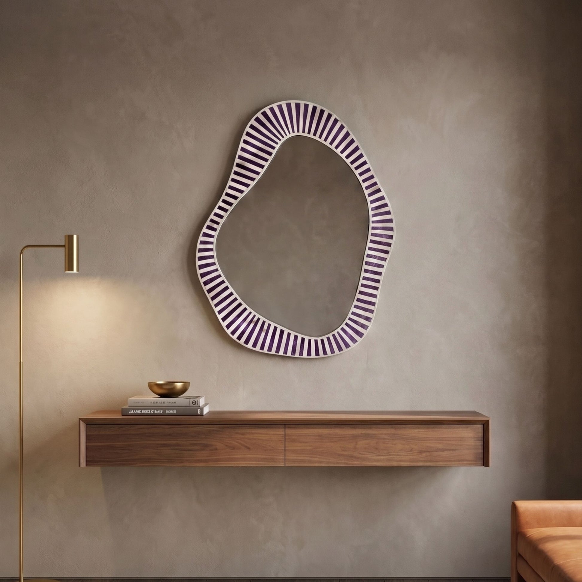 An organic-shaped wall mirror with a purple and white striped frame hangs on a textured beige wall above a floating wooden console. A brass floor lamp stands to the left, and a small decorative bowl and books sit on the shelf.