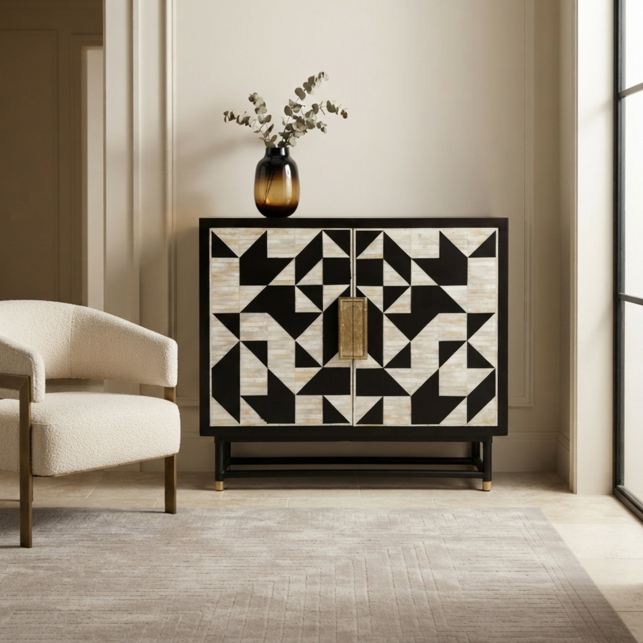 A modern two-door sideboard or credenza with a bold black and off-white geometric inlay pattern, featuring a central gold-toned rectangular handle and gold feet. It is styled in a minimalist room with a cream armchair, a vase of dried branches, and a textured rug.