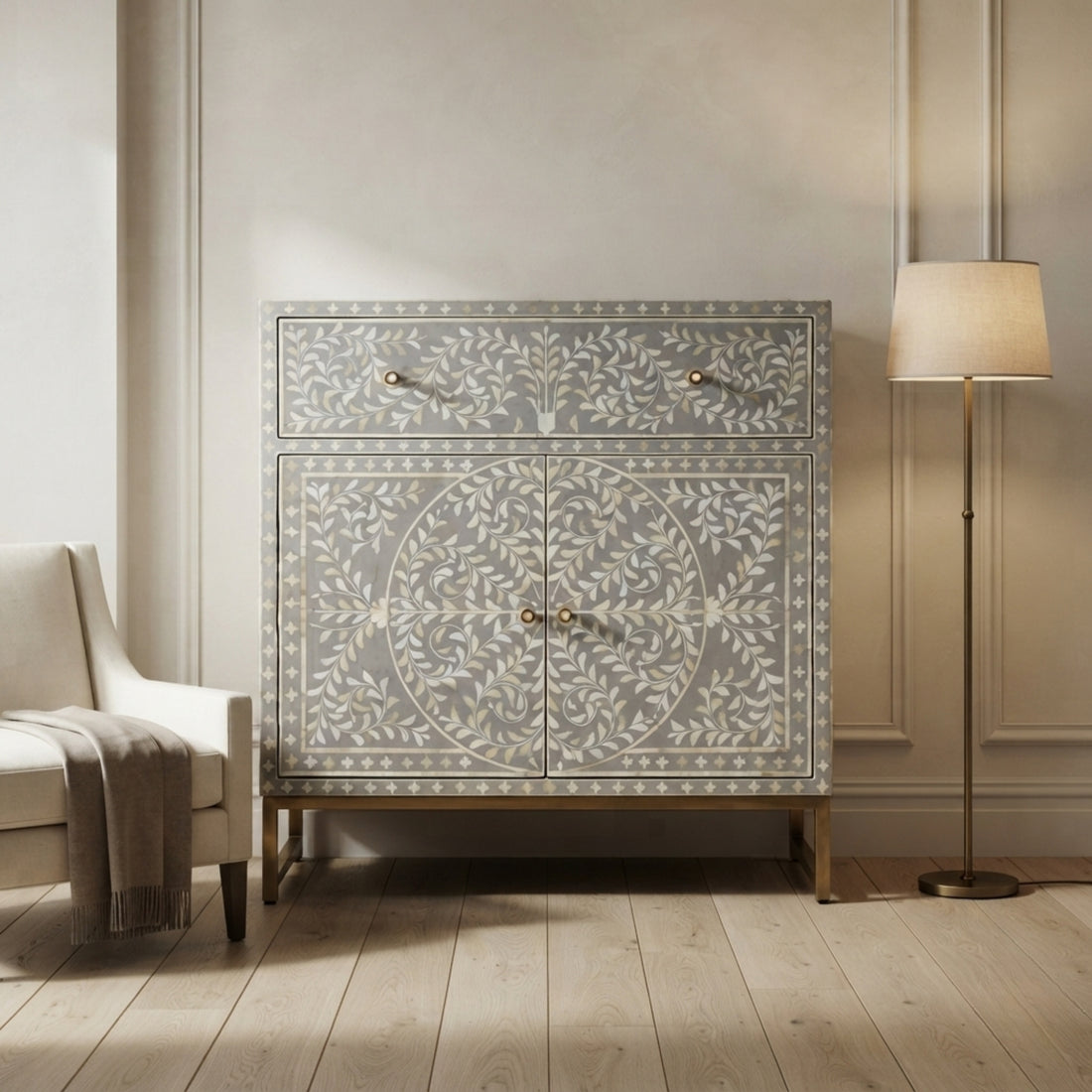 A tall decorative cabinet with two upper drawers and two lower doors, featuring an intricate ivory inlay pattern on a light gray wood base. The cabinet stands in a styled room with a light wood floor, a cream armchair on the left, and a floor lamp on the right.