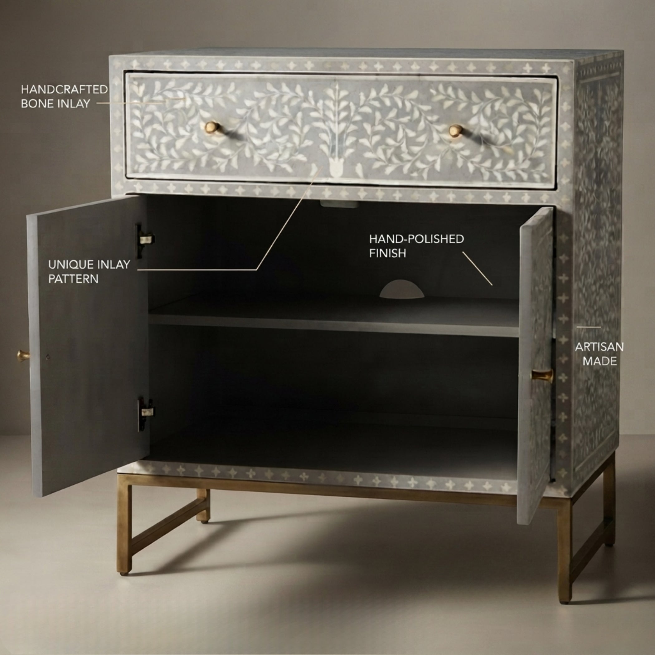 A handcrafted two-drawer cabinet with open doors, featuring a light gray bone-inlay pattern with leaf motifs, gold-tone knobs, and brass legs. White text labels point out key features like handcrafted bone inlay, unique inlay pattern, hand-polished finish, and artisan made, set against a neutral studio background.