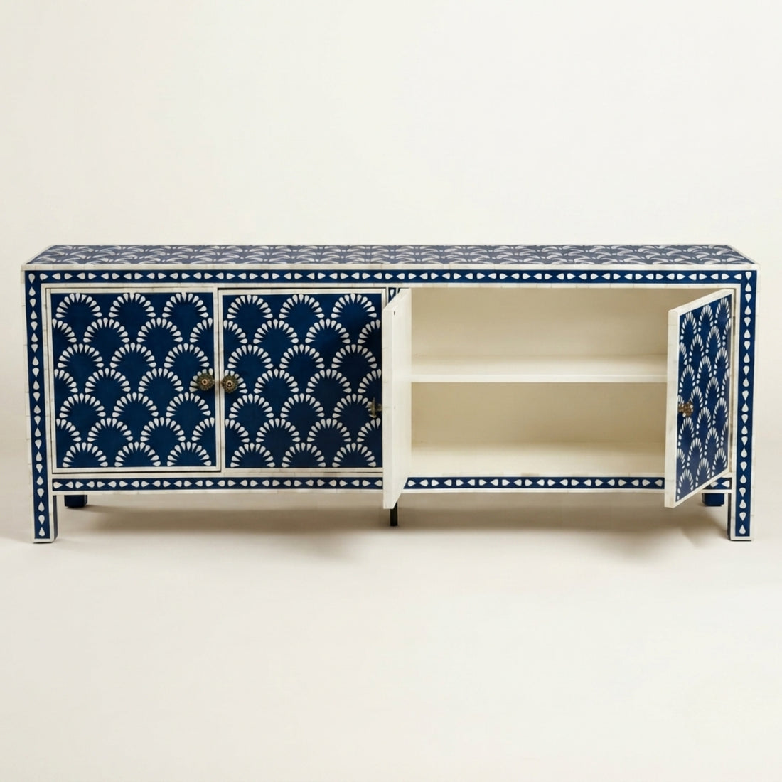 A blue and white patterned sideboard cabinet in a modern, warm-toned room with a textured beige wall, wooden floor, and an open door showing a garden view. The cabinet has three doors, with the center door open to reveal shelves, and is styled with two table lamps and a small sculpture.