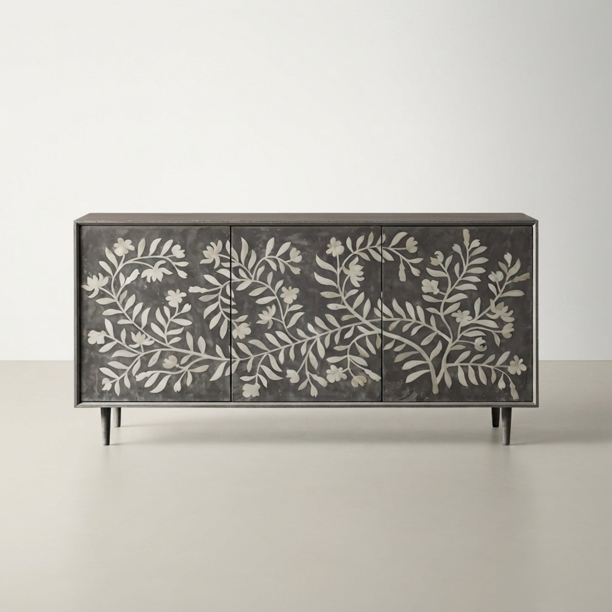 A rectangular sideboard or credenza with three doors, featuring a dark charcoal gray finish and intricate white floral and leaf patterns across the front. The cabinet has slim black legs and is photographed against a neutral, light gray studio background.