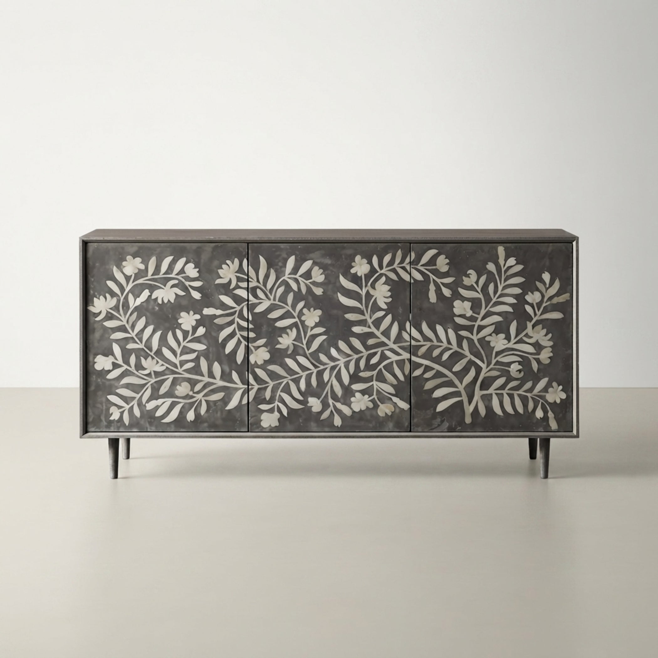 A rectangular sideboard or credenza with three doors, featuring a dark charcoal gray finish and intricate white floral and leaf patterns across the front. The cabinet has slim black legs and is photographed against a neutral, light gray studio background.