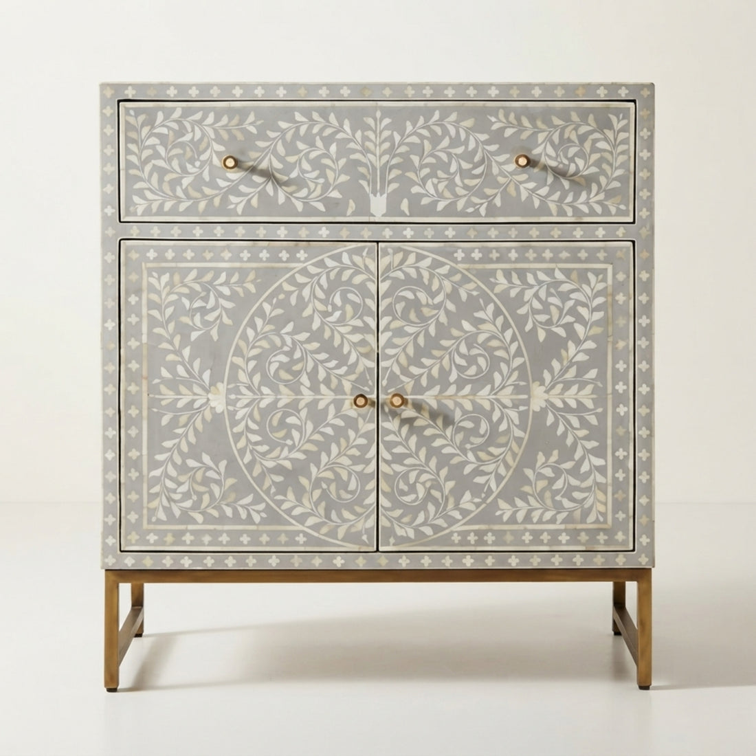 A tall decorative cabinet with two upper drawers and two lower doors, featuring an intricate ivory inlay pattern on a light gray wood base. The cabinet stands in a styled room with a light wood floor, a cream armchair on the left, and a floor lamp on the right.