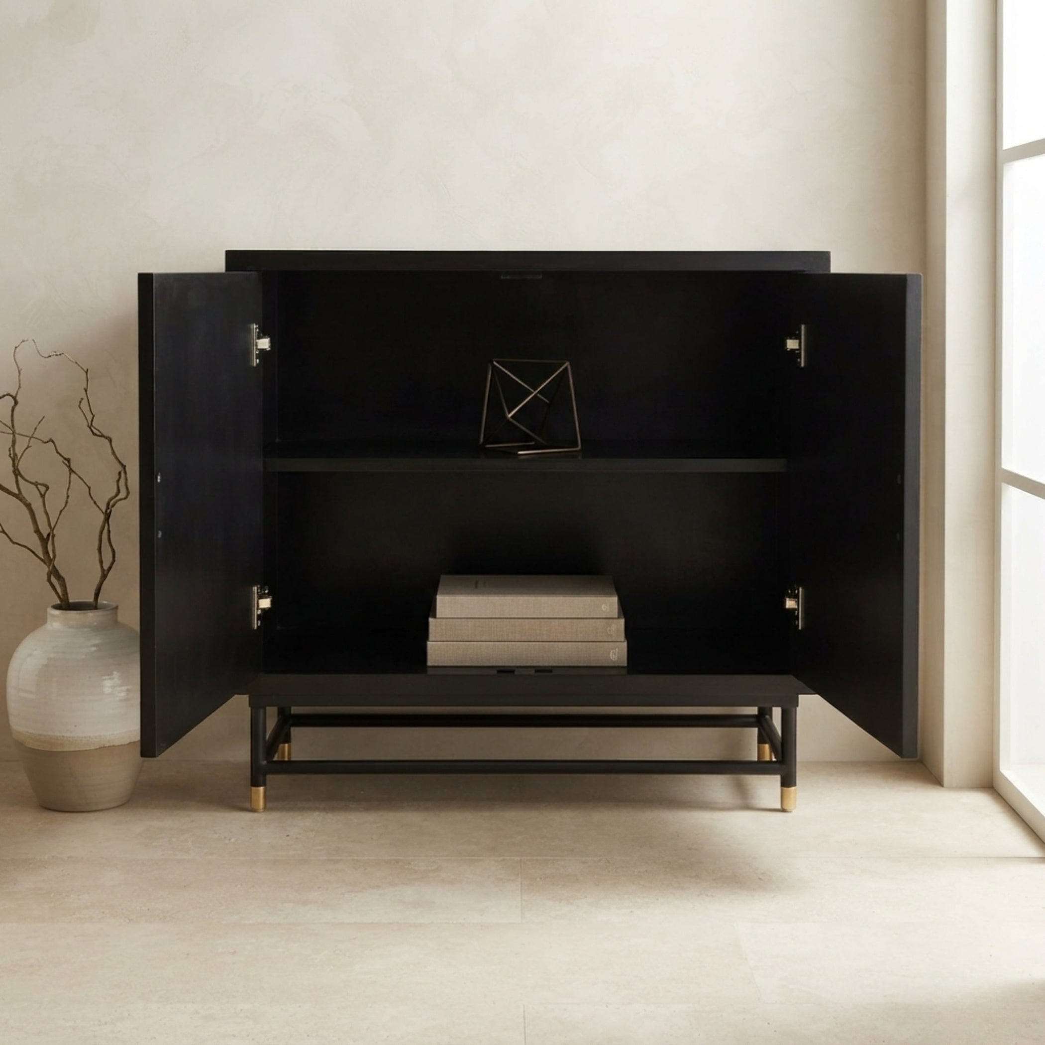 A modern black sideboard cabinet with open doors, styled with books and a geometric metal vase on a shelf, set against a neutral beige wall and floor.