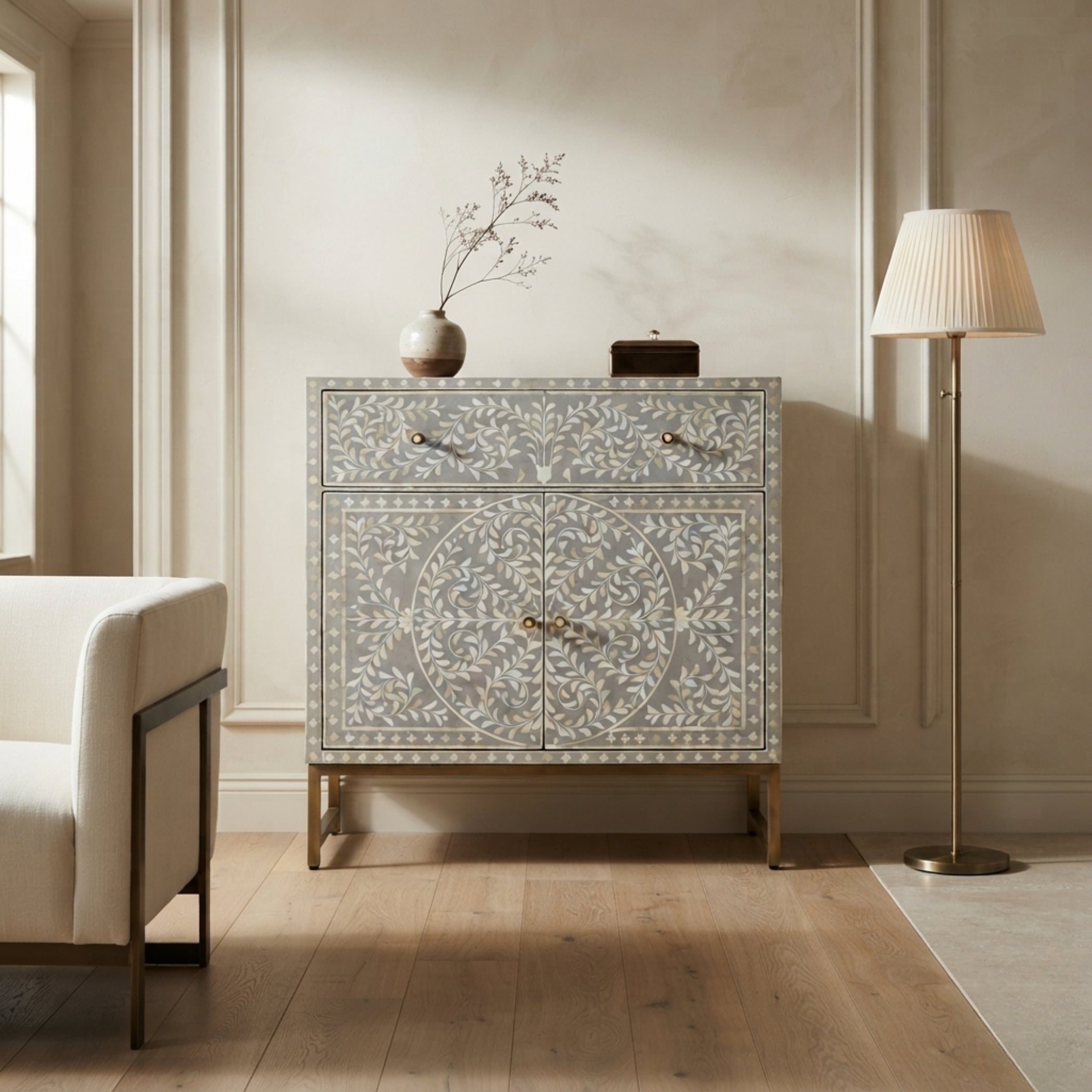 A light gray bone-inlay chest of drawers with gold legs, styled in a warm beige room next to a white armchair and a floor lamp.