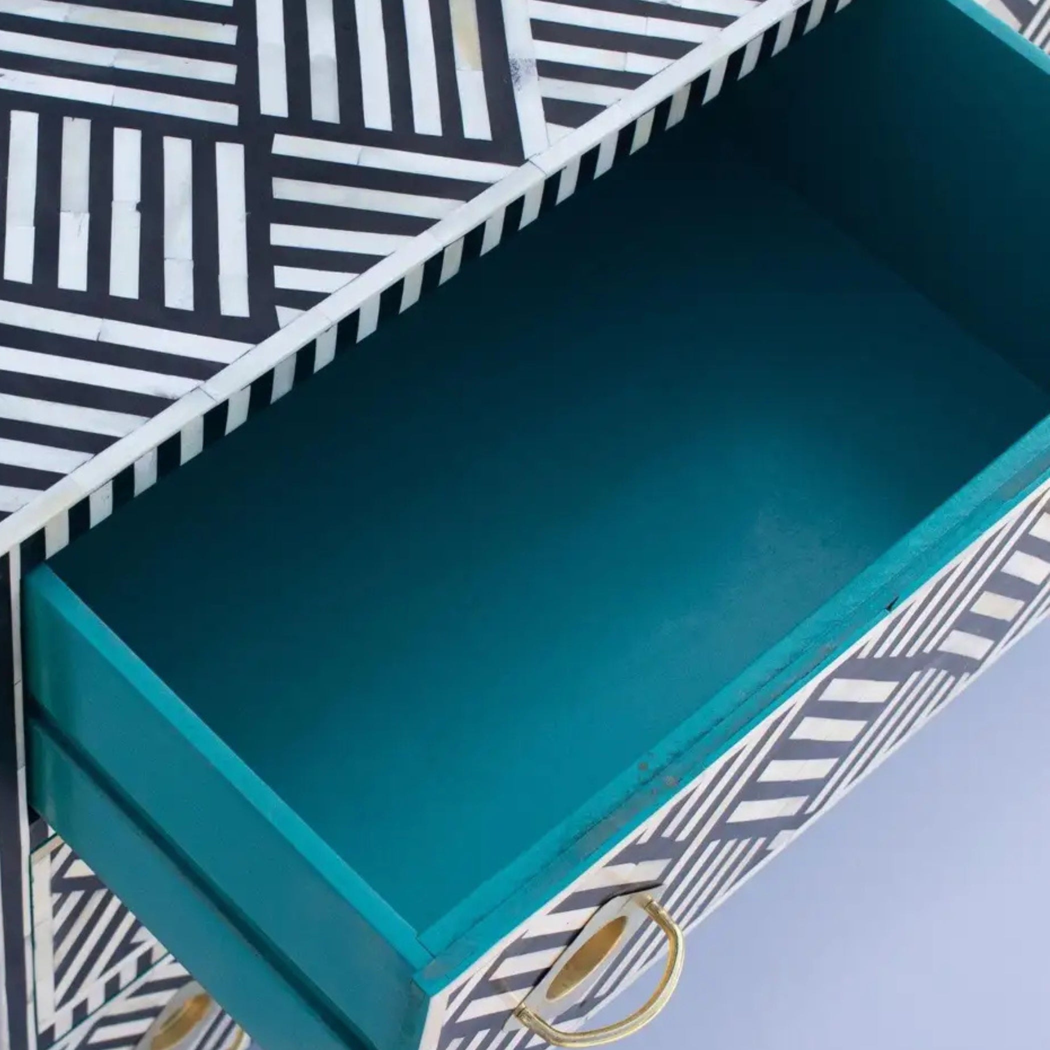 Close-up of an open drawer with a teal interior, set in a piece of furniture featuring a black and white geometric striped pattern on the exterior, with a gold pull handle.
