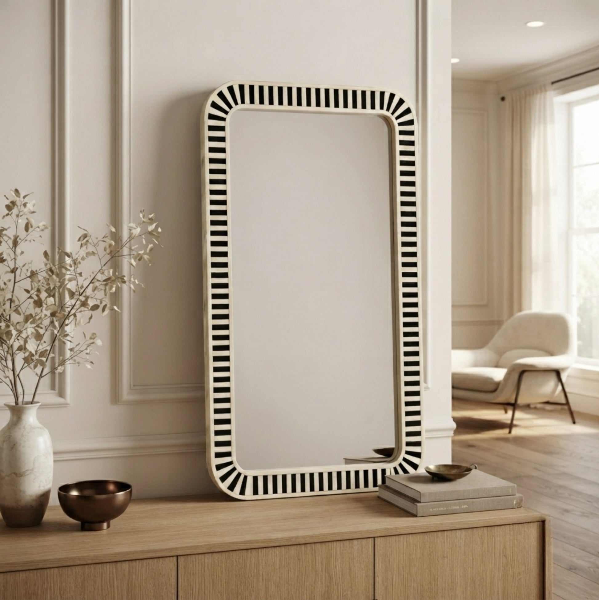A tall rectangular wall mirror with a black-and-white vertically striped border, leaning against a cream paneled wall on a light wood console in a styled interior room.