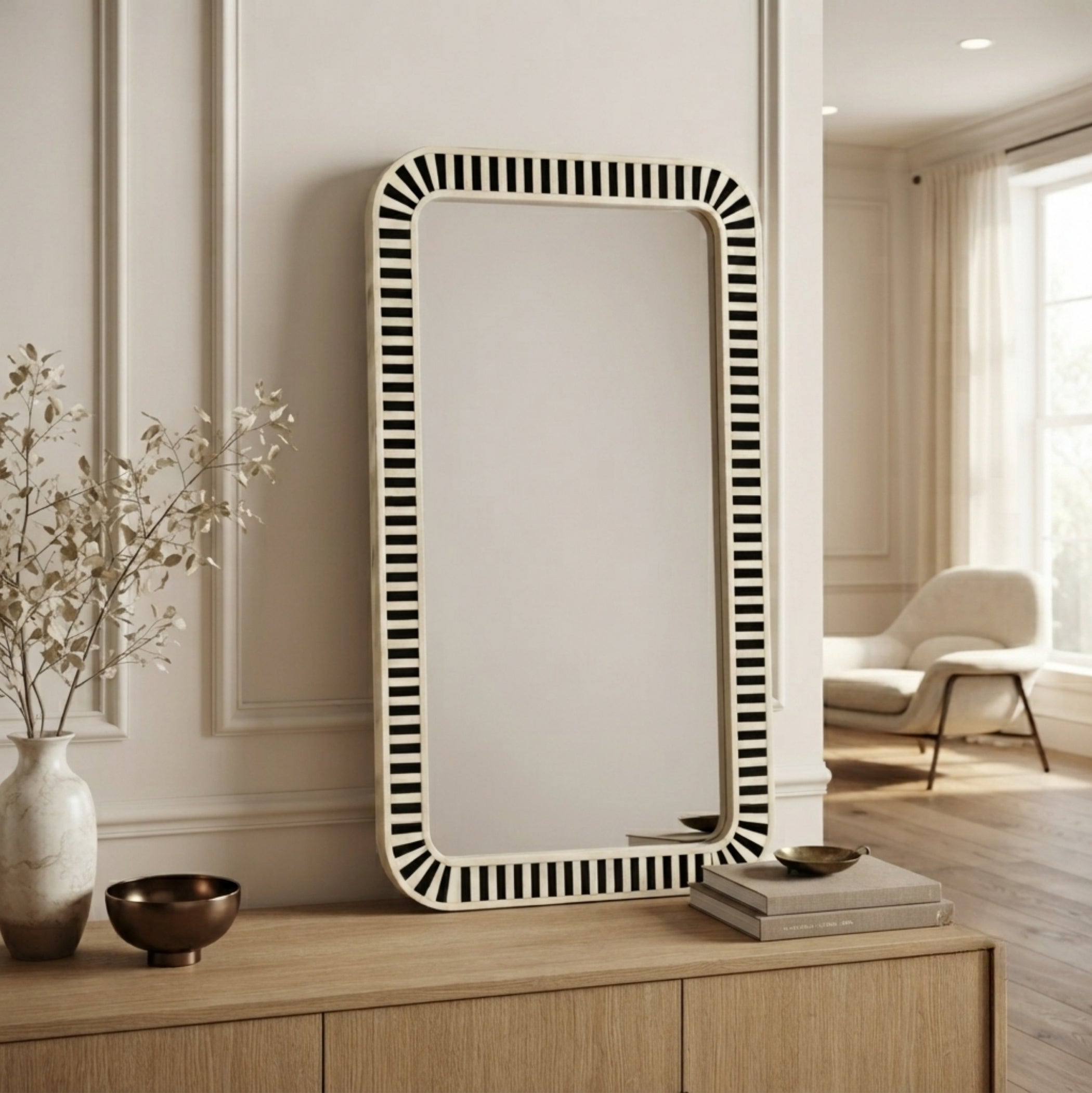 A tall rectangular wall mirror with a black-and-white vertically striped border, leaning against a cream paneled wall on a light wood console in a styled interior room.