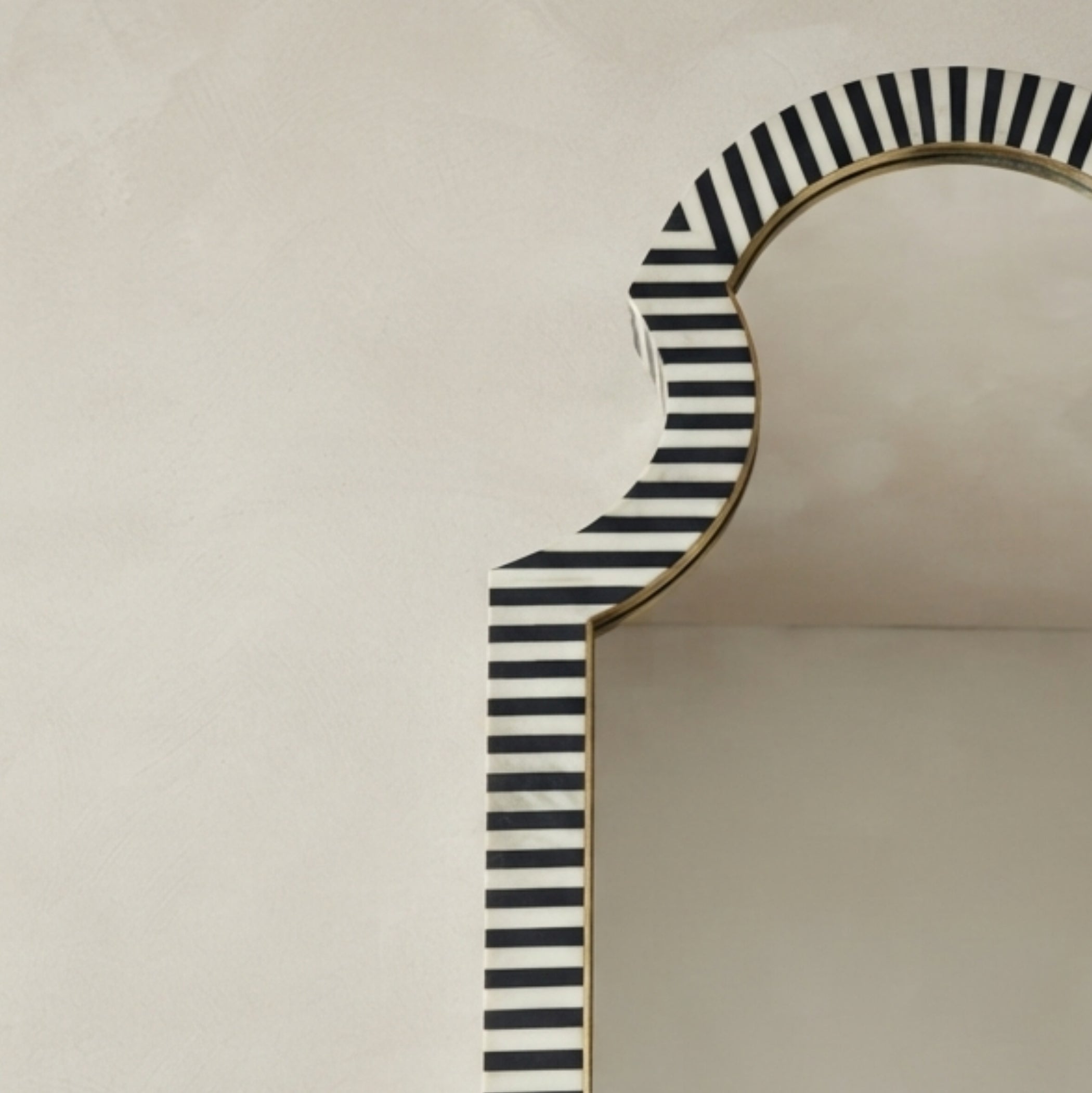 A close-up of a decorative arch-shaped mirror with a black and white striped pattern and a gold-toned frame, set against a warm beige textured wall.