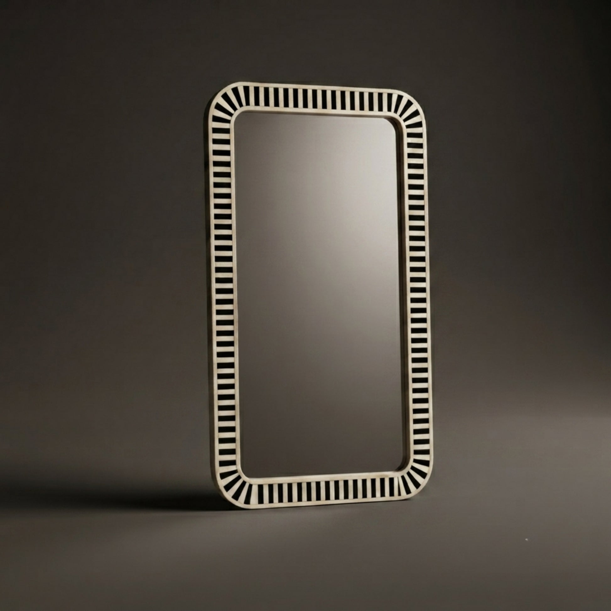 A rectangular wall mirror with a decorative off-white frame featuring a repeating pattern of small vertical slits, standing on a dark gray surface against a dark gray background.