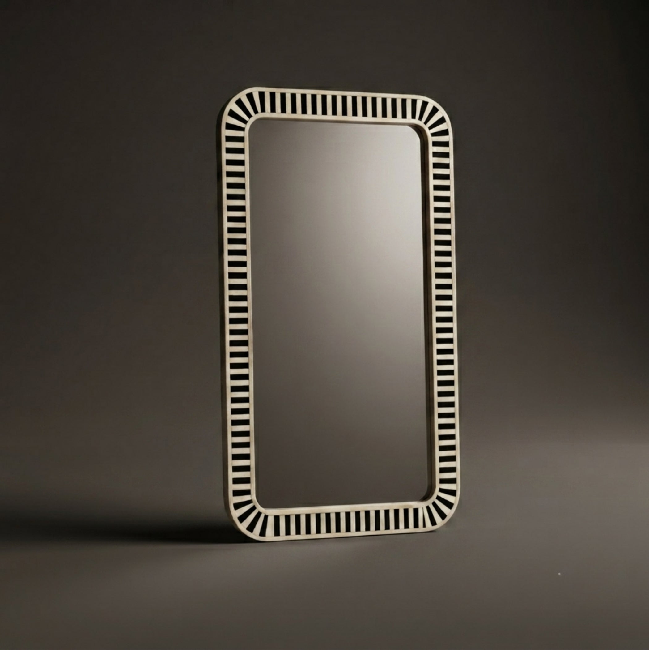 A rectangular wall mirror with a decorative off-white frame featuring a repeating pattern of small vertical slits, standing on a dark gray surface against a dark gray background.