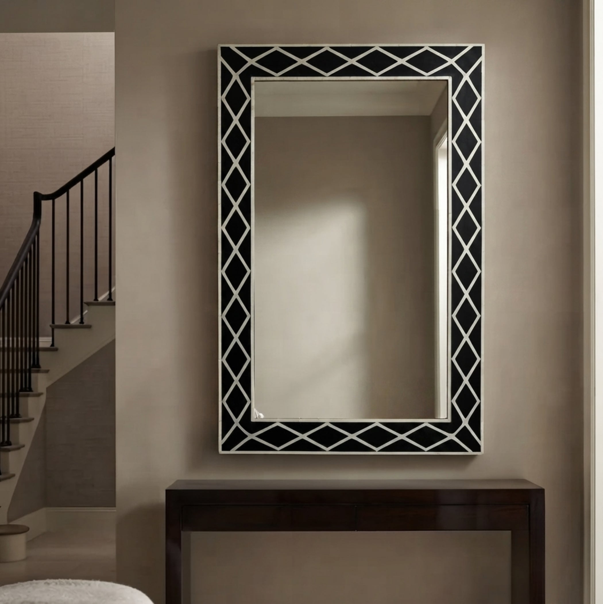 A rectangular wall mirror with a black and white geometric lattice border, mounted on a beige wall above a dark wood console table, with a staircase visible in the background.