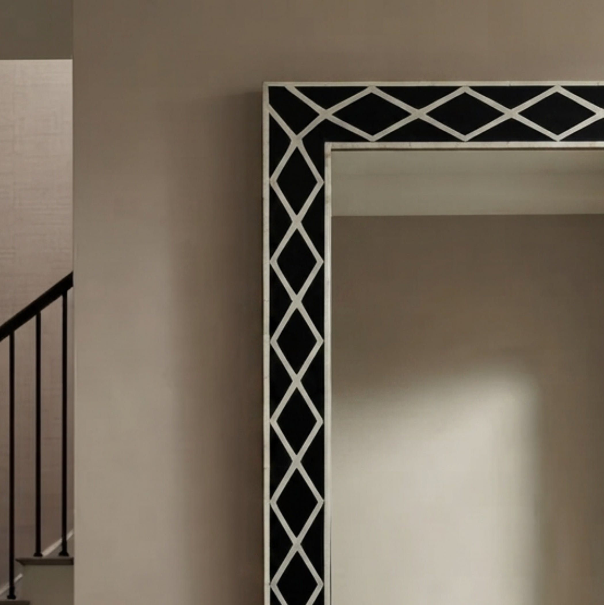 A large rectangular mirror with a black and white geometric diamond-patterned frame, shown mounted on a beige wall in an interior setting with a staircase visible on the left.