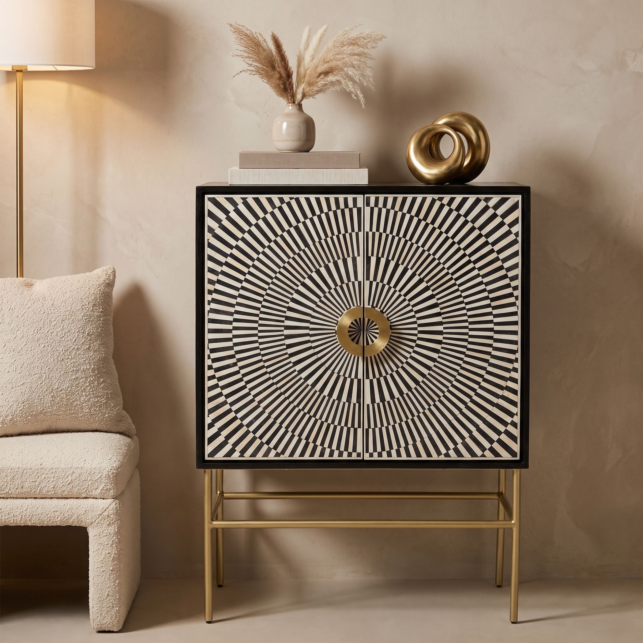 A modern sideboard cabinet with a black and white geometric sunburst pattern on the doors and a gold metal frame and legs, styled in a warm beige room next to a textured armchair.