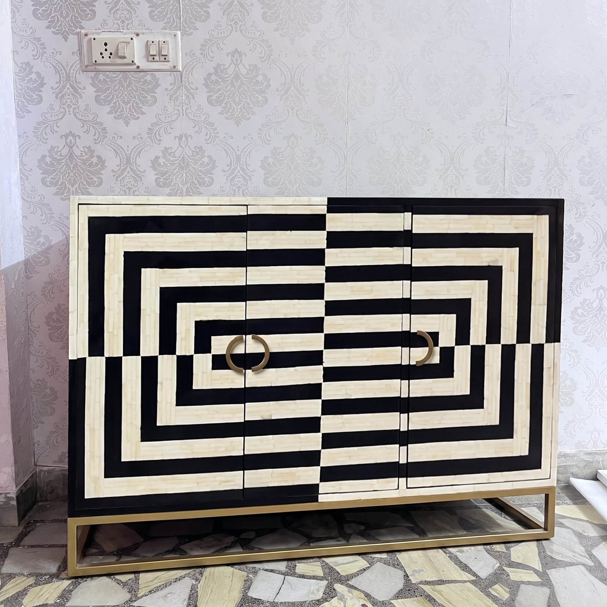 A modern sideboard cabinet with a bold black and cream geometric striped pattern, featuring two circular gold handles and a gold metal base, set against a patterned wallpaper background.