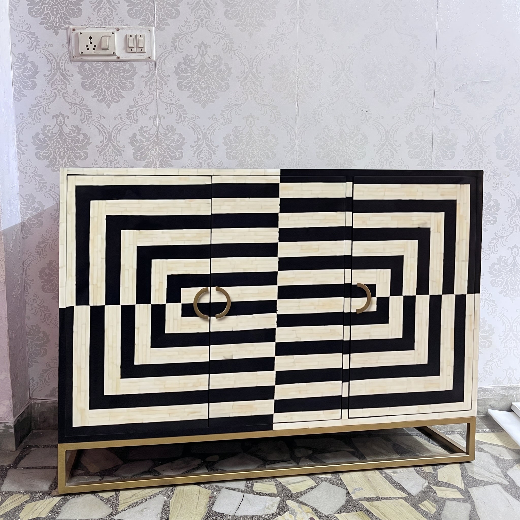 A modern sideboard cabinet with a bold black and cream geometric striped pattern, featuring two circular gold handles and a gold metal base, set against a patterned wallpaper background.