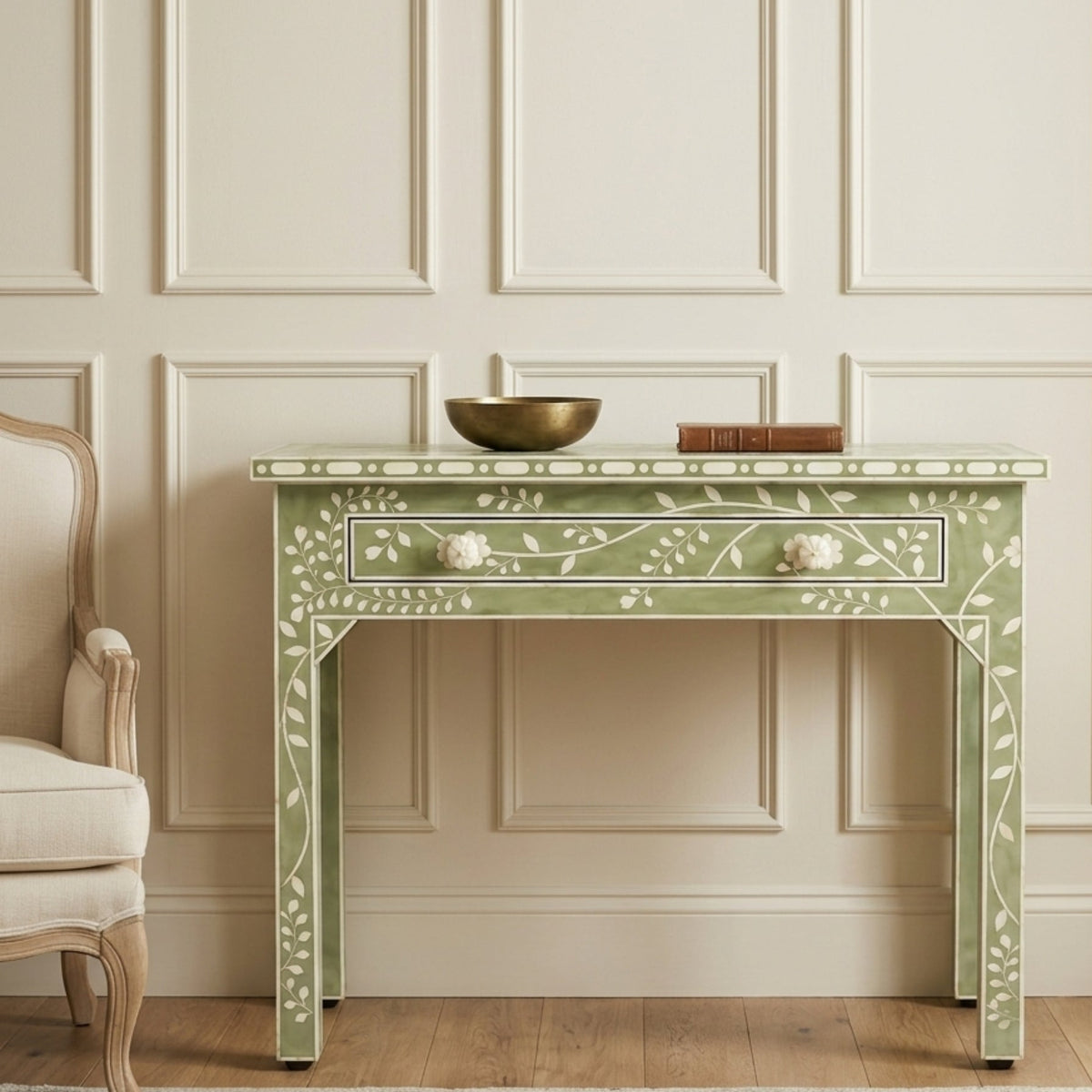 A light sage green console table with white floral inlay and a single drawer, styled in a classic room with a cream paneled wall, wood floor, and a partial cream armchair.
