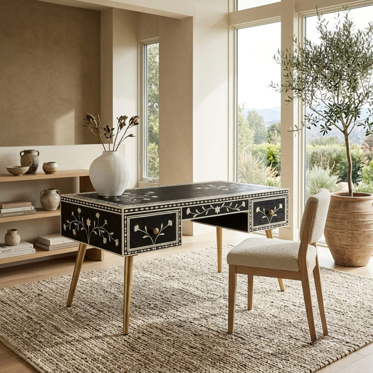 A black writing desk with gold legs and white floral inlay, styled in a bright, modern room with a matching upholstered chair, a large potted tree, and a vase on top.