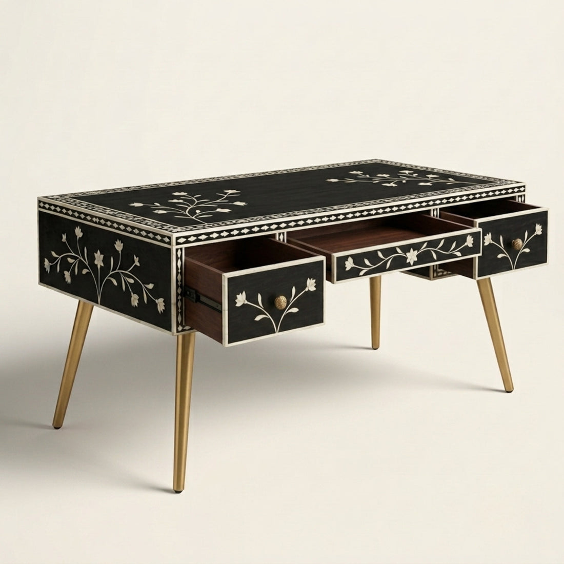 A black writing desk with gold legs and white floral inlay, styled in a bright, modern room with a matching upholstered chair, a large potted tree, and a vase on top.