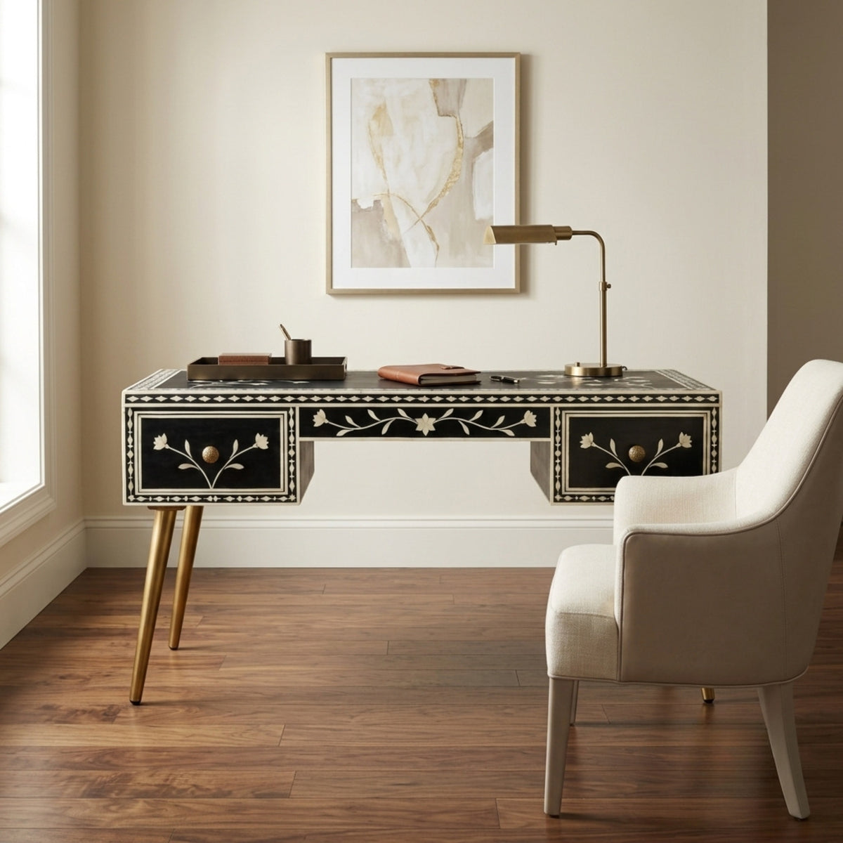 A black and gold desk with floral inlay details and gold legs, styled in a home office setting next to a cream-colored armchair and under a framed abstract print.
