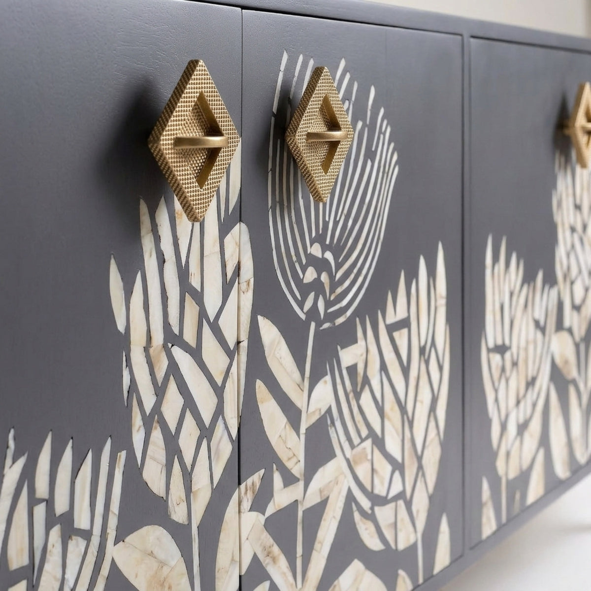 A close-up of a dark gray wooden cabinet featuring a cream-colored inlay of stylized floral and leaf motifs, with two prominent textured gold diamond-shaped drawer pulls.