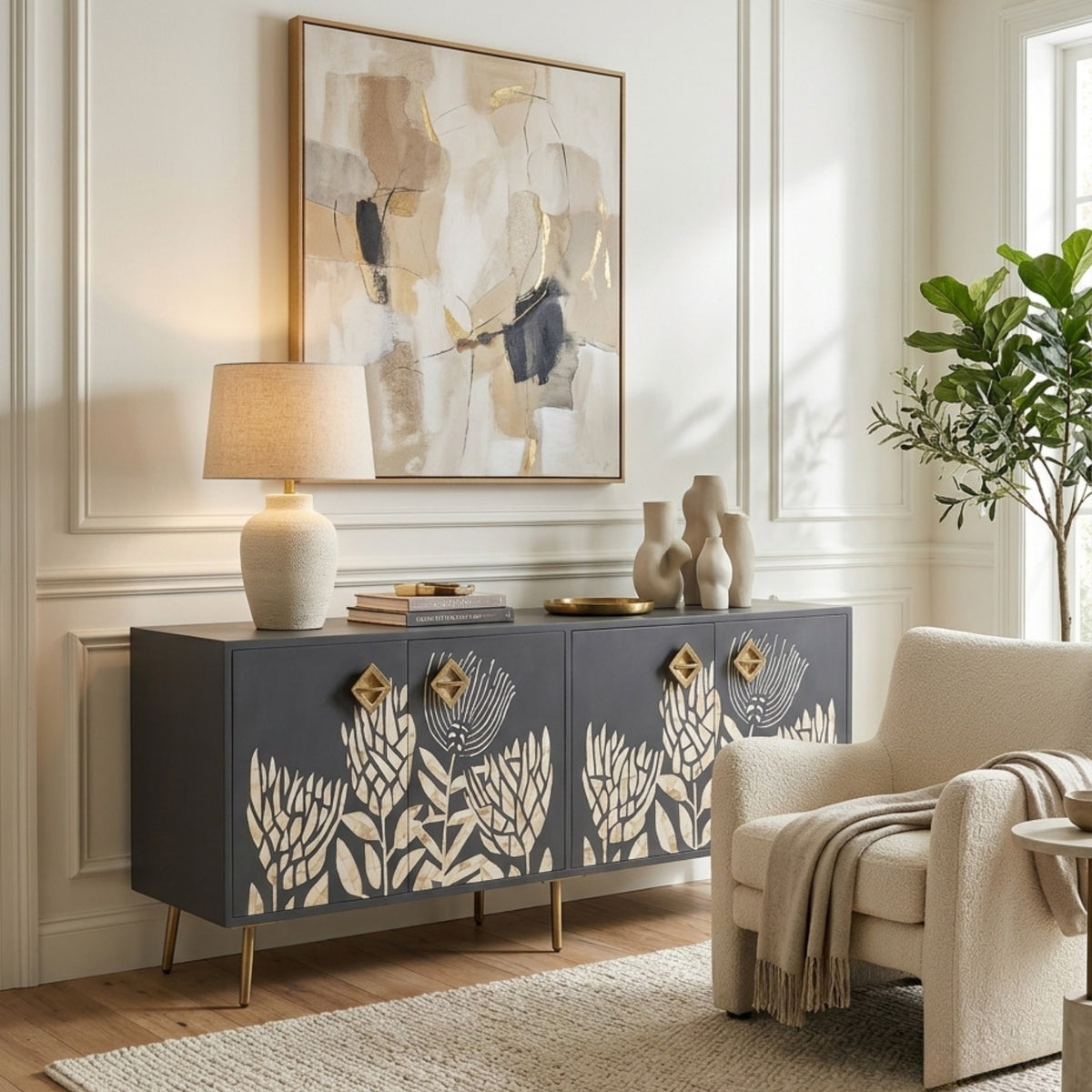 A dark gray sideboard cabinet with cream floral cutout panels and gold legs, styled in a bright living room next to a beige armchair and under a framed abstract painting.