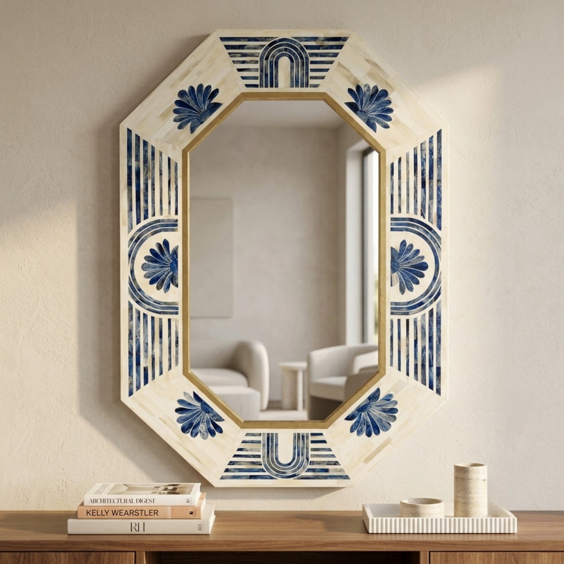 An octagonal wall mirror with a cream-colored distressed frame featuring blue floral and geometric patterns, mounted on a textured beige wall above a wooden console table with books and decor.