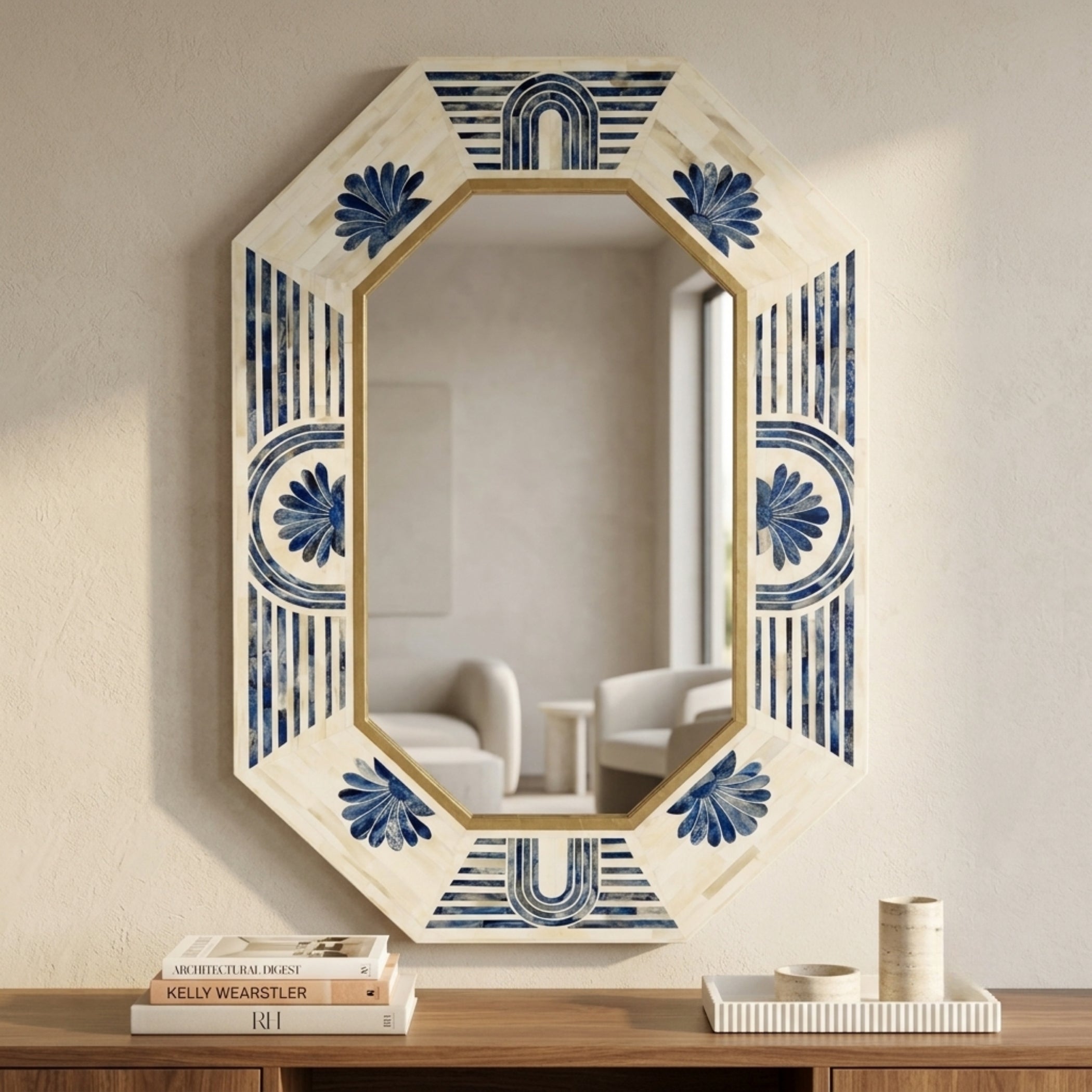 An octagonal wall mirror with a cream-colored distressed frame featuring blue floral and geometric patterns, mounted on a textured beige wall above a wooden console table with books and decor.