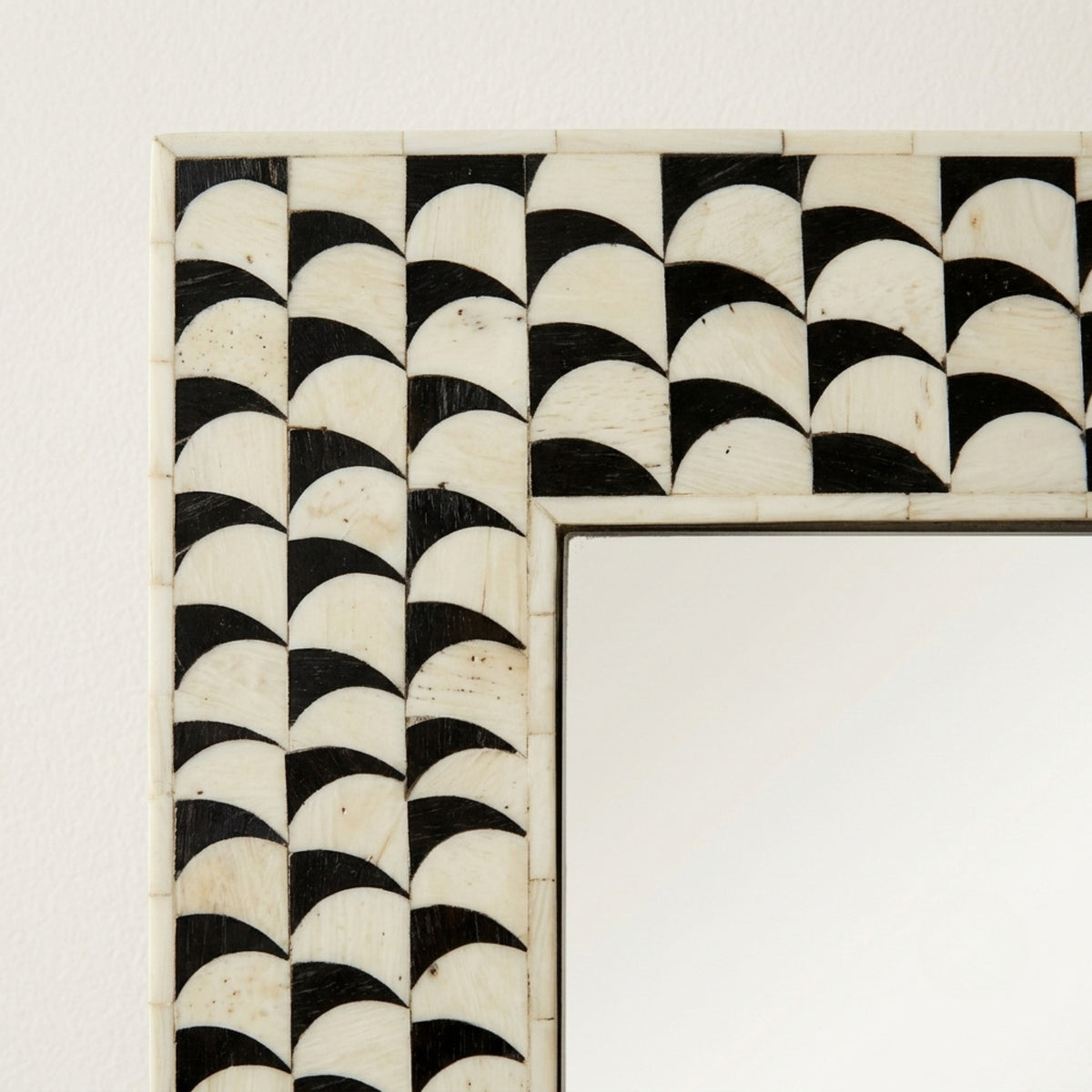 A close-up of a rectangular mirror frame featuring a black and off-white scalloped geometric pattern, mounted on a light-colored wall.