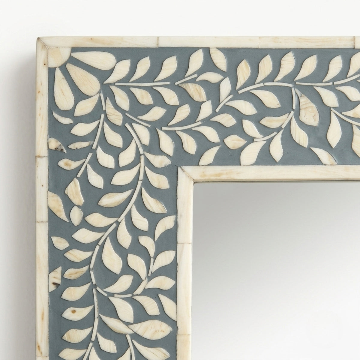 A close-up of a decorative rectangular frame with an intricate inlay pattern of cream-colored leaf shapes over a muted blue-gray background, set against a plain white wall.