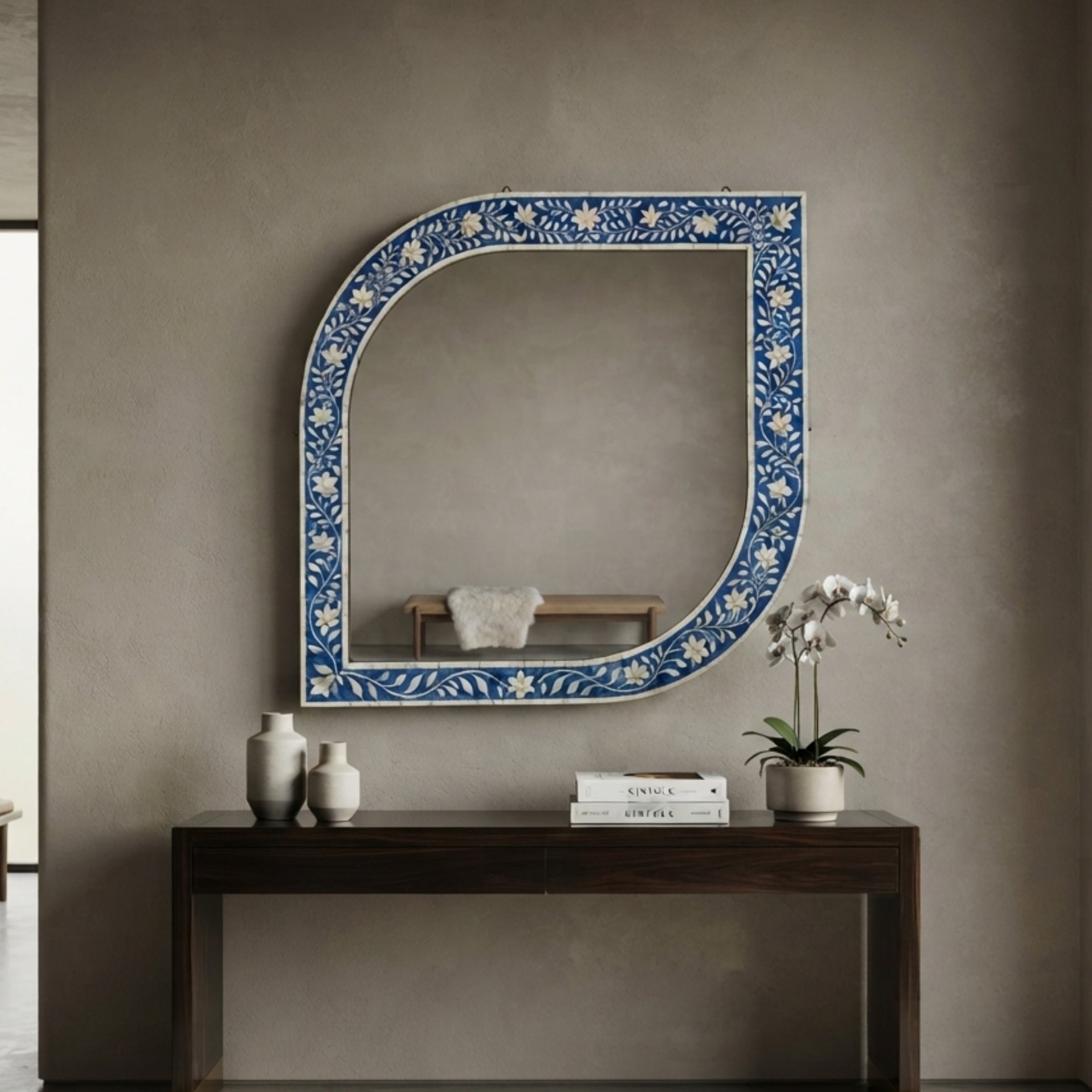 An arched decorative wall mirror with a blue and white floral patterned border, mounted on a textured taupe wall above a dark wood console table with vases, books, and a potted orchid.