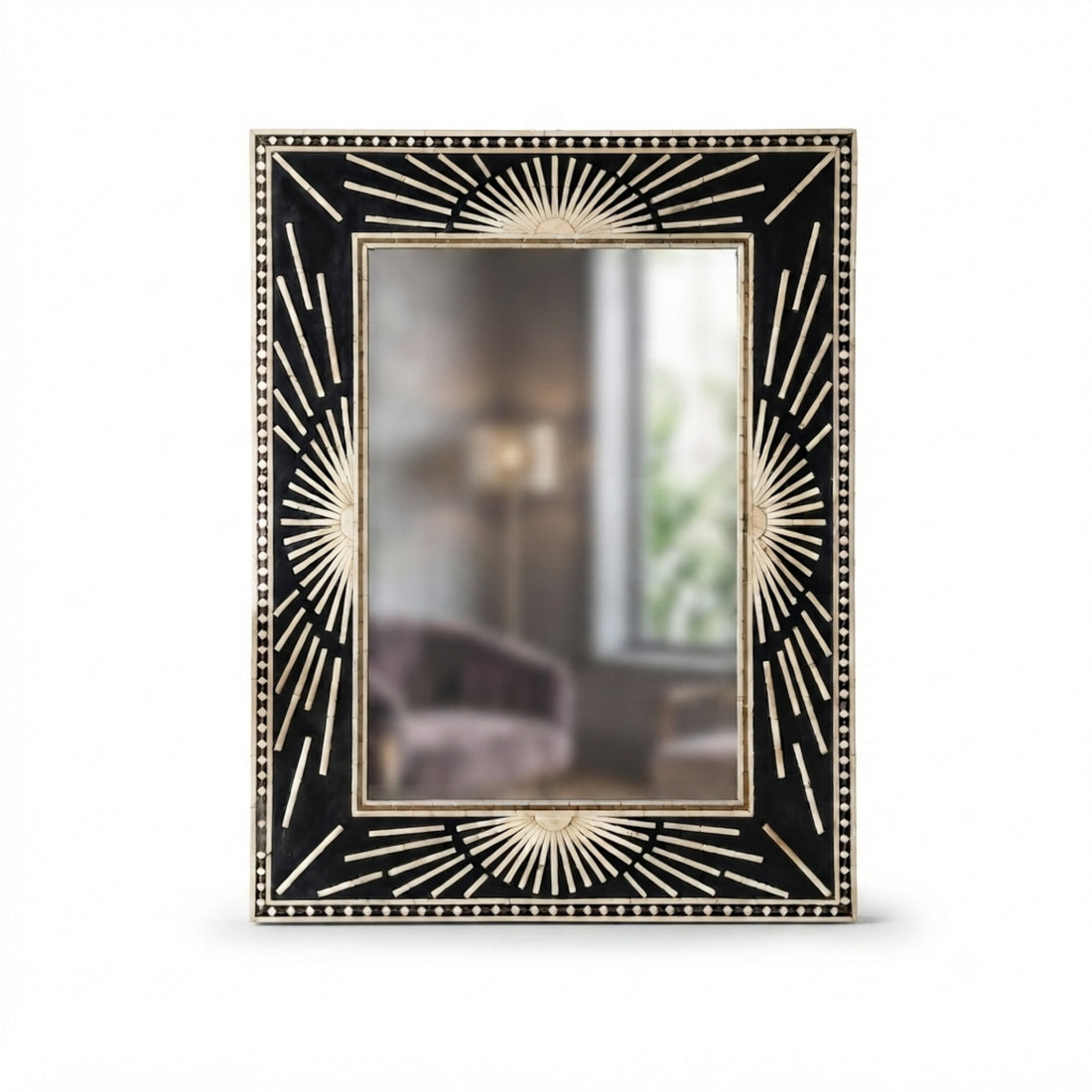 A large rectangular wall mirror with an ornate black and gold sunburst frame hangs above a dark wood console table in a styled room. A white ceramic vase with dried branches, stacked books, a table lamp, and a round textured ottoman with a draped brown throw are arranged below.