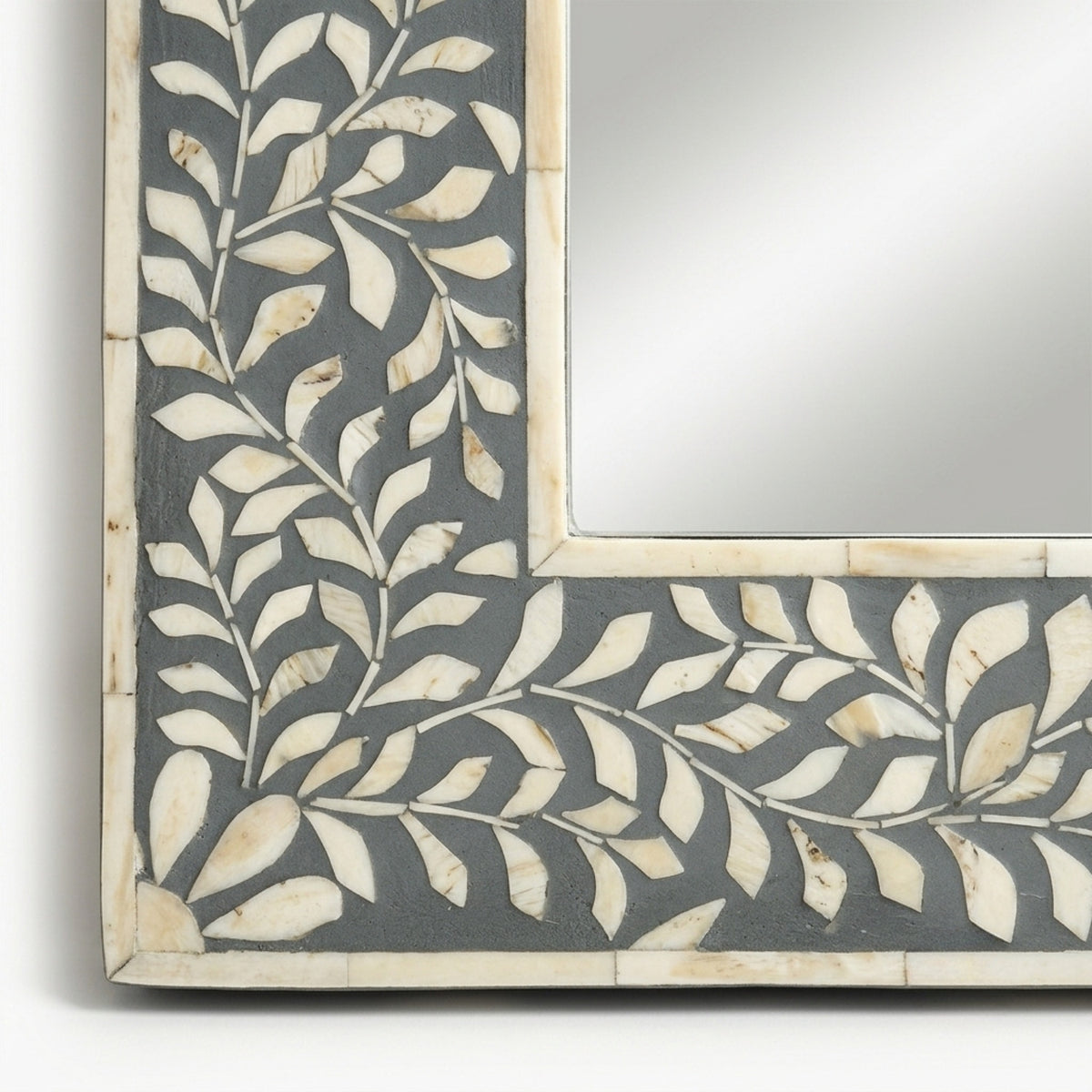 A close-up of a decorative square mirror frame featuring an inlay pattern of cream-colored leaf shapes over a dark charcoal gray background.