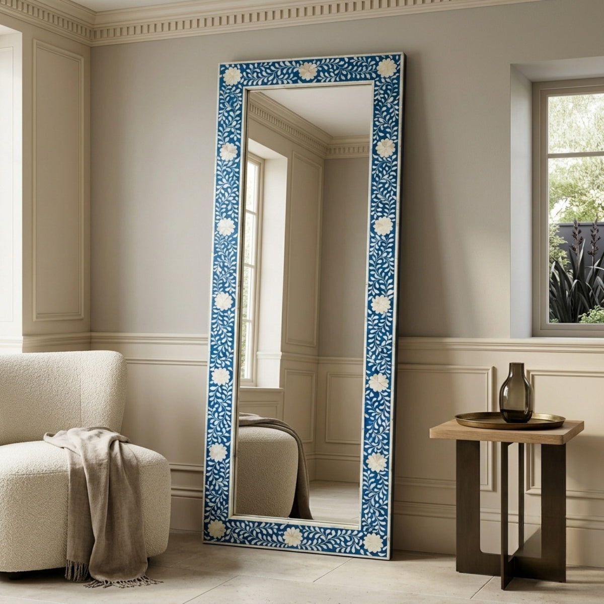 A tall floor mirror with a blue and white floral patterned frame, leaning against a beige paneled wall in a styled room with a chair and side table.