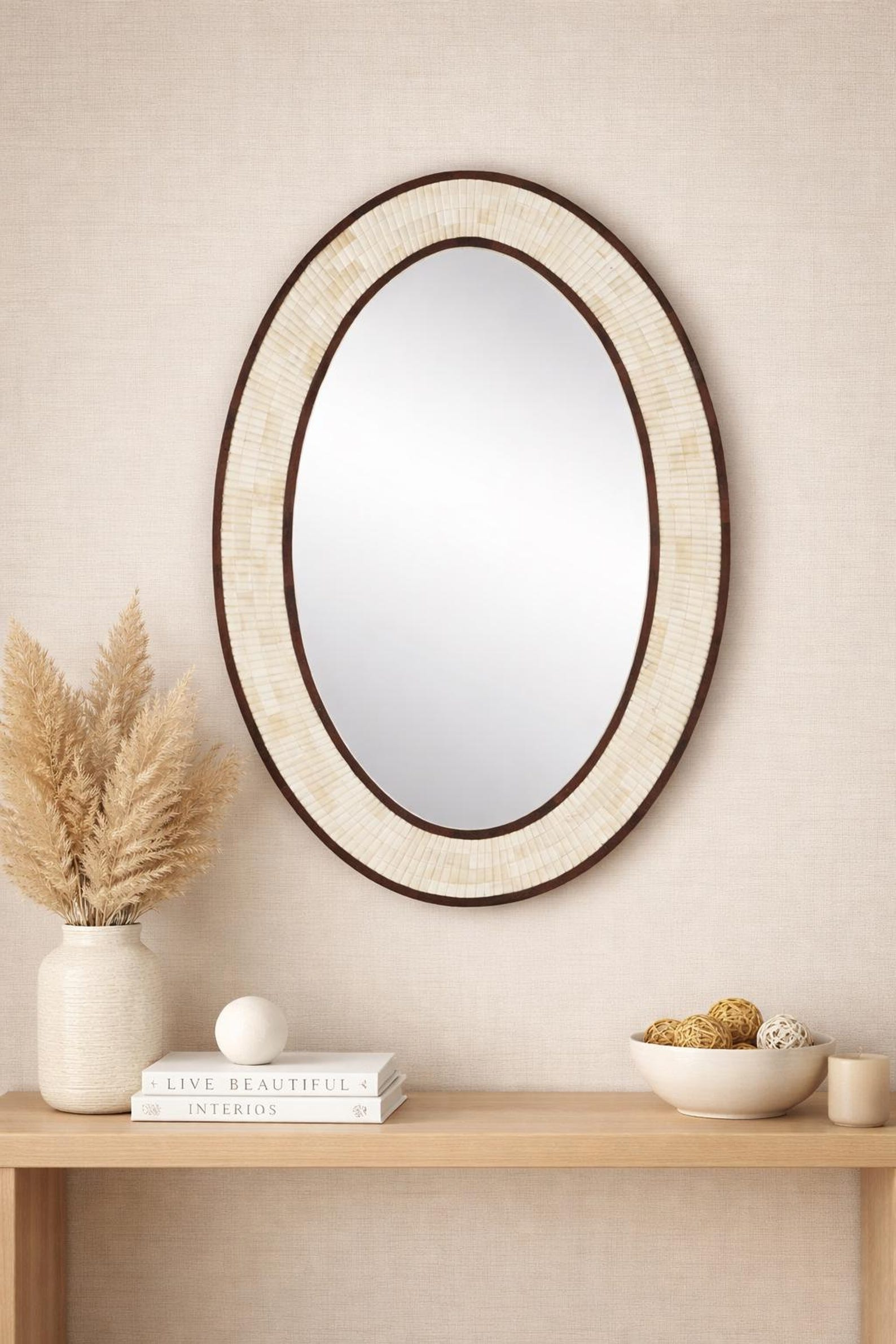 An oval wall mirror with a textured cream and beige frame and a dark brown border, styled on a light wood console table against a beige wall with decorative props.